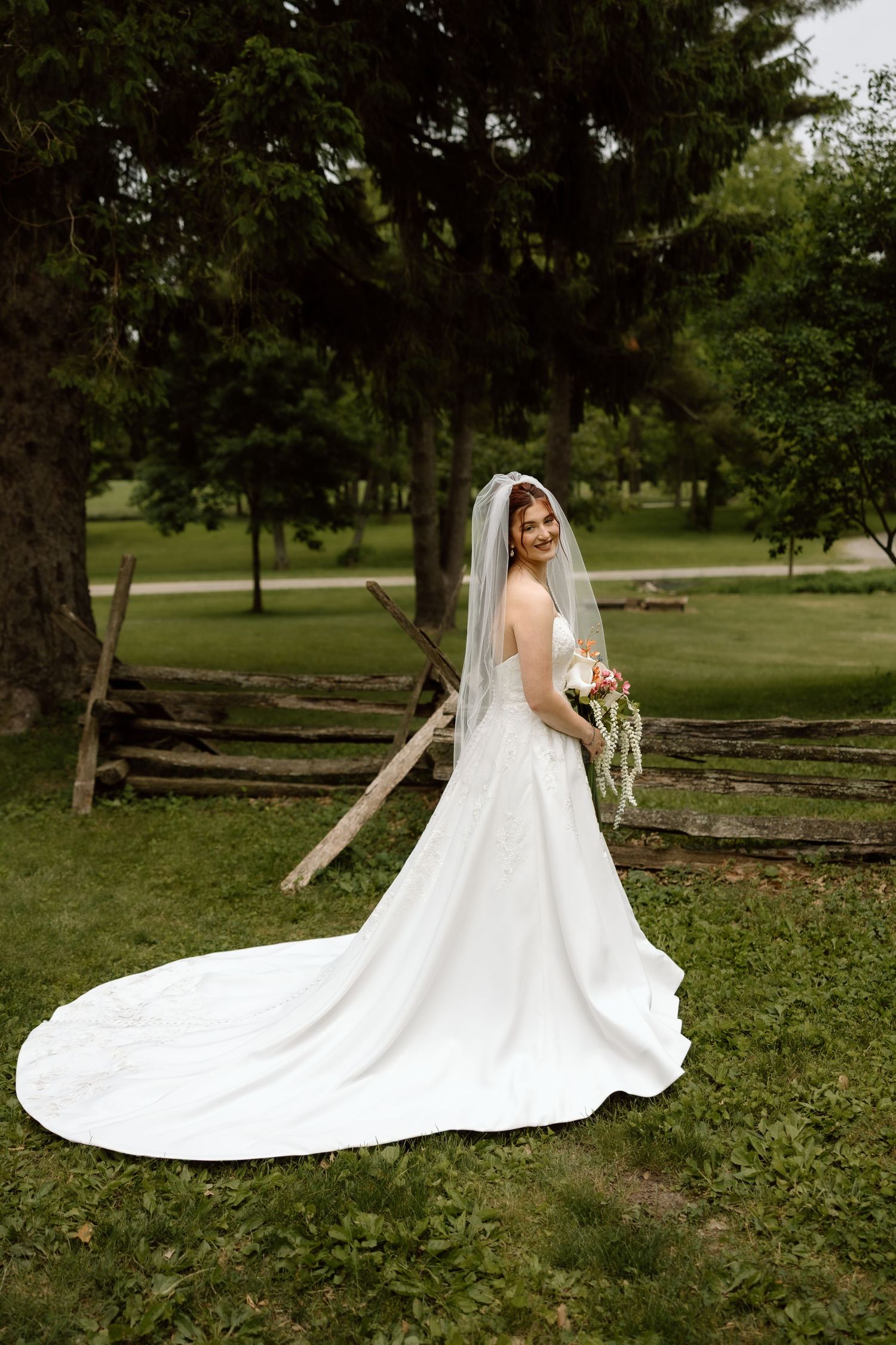 Smiling bride in strapless white gown with long train and veil poses outdoors near rustic fence.
