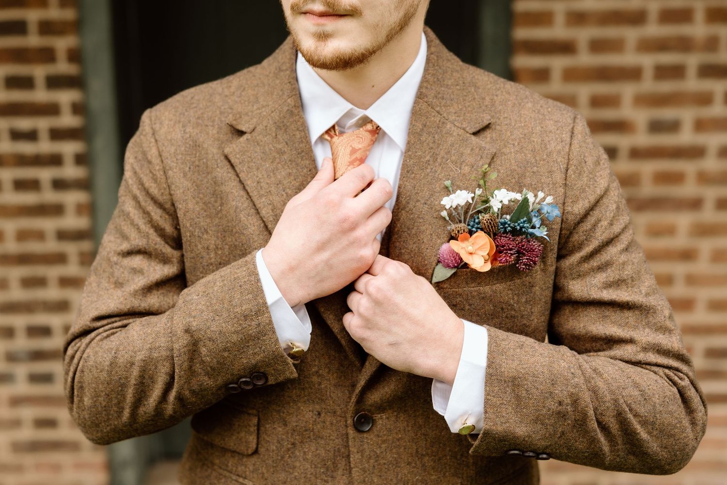 Groom in brown tweed suit adjusting orange tie with colorful floral boutonniere on lapel.