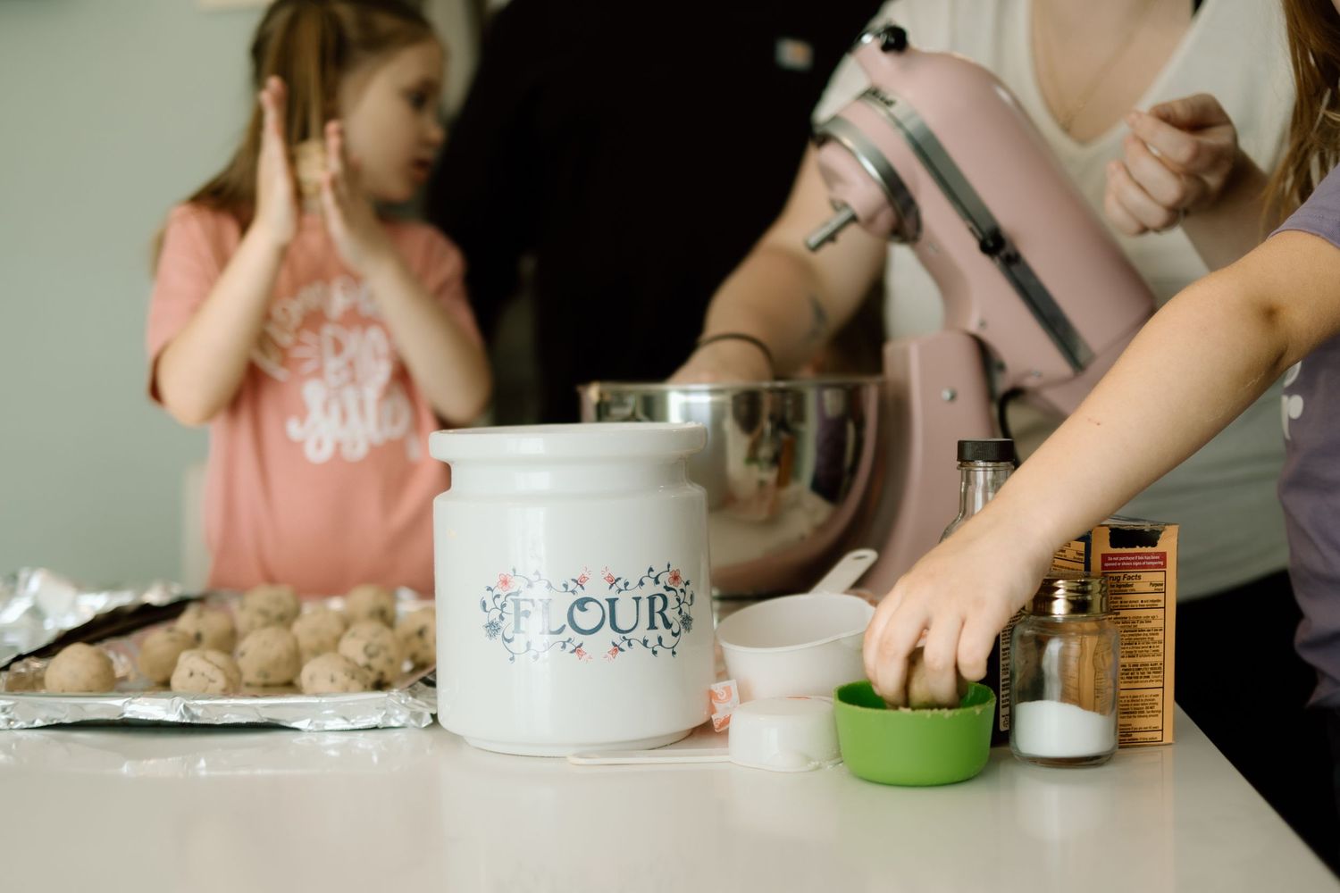 Children and mother baking cookies with a pink KitchenAid mixer and white flour canister on counter.