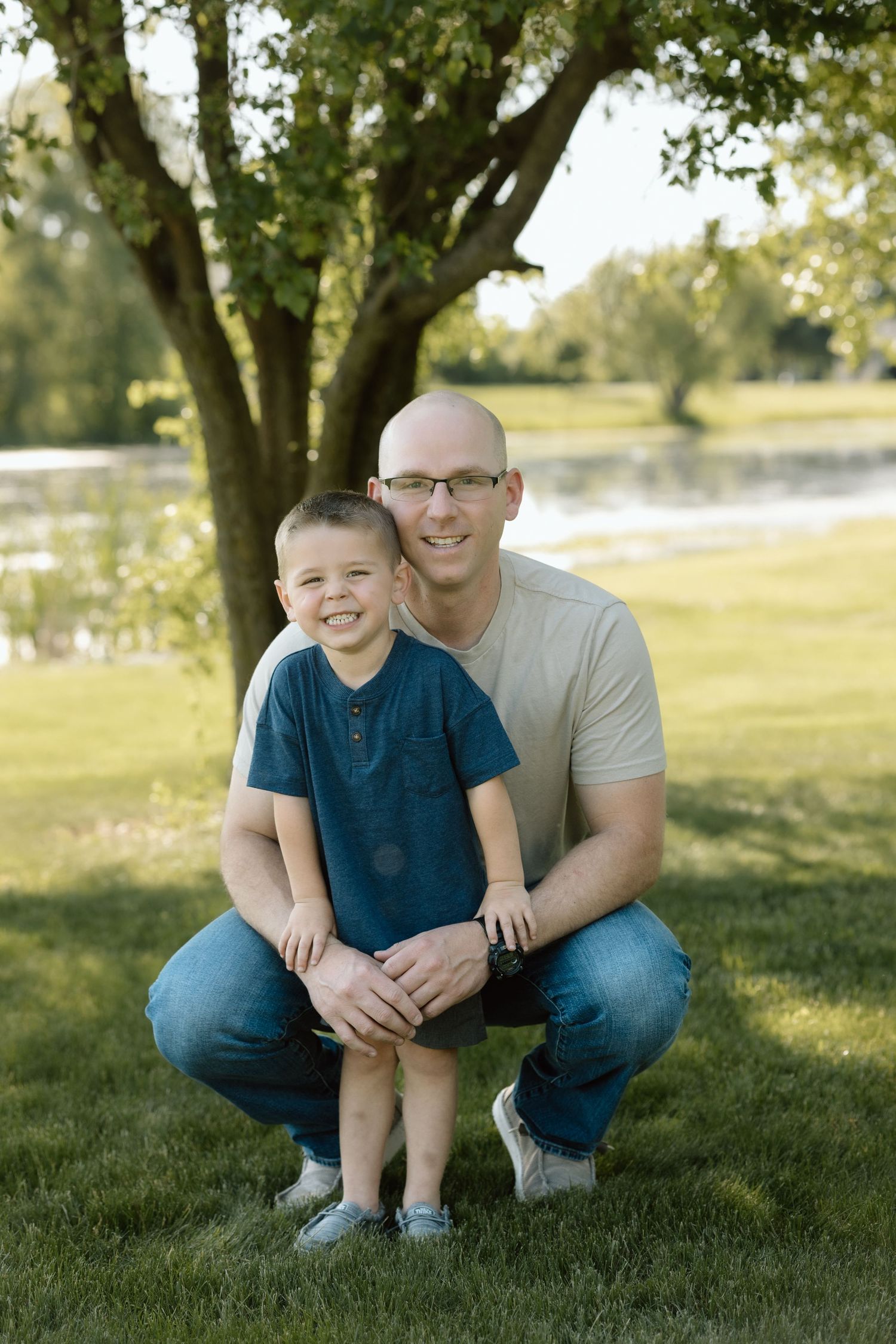 Bald father with glasses crouches beside smiling young son in blue shirt for outdoor family portrait near pond.