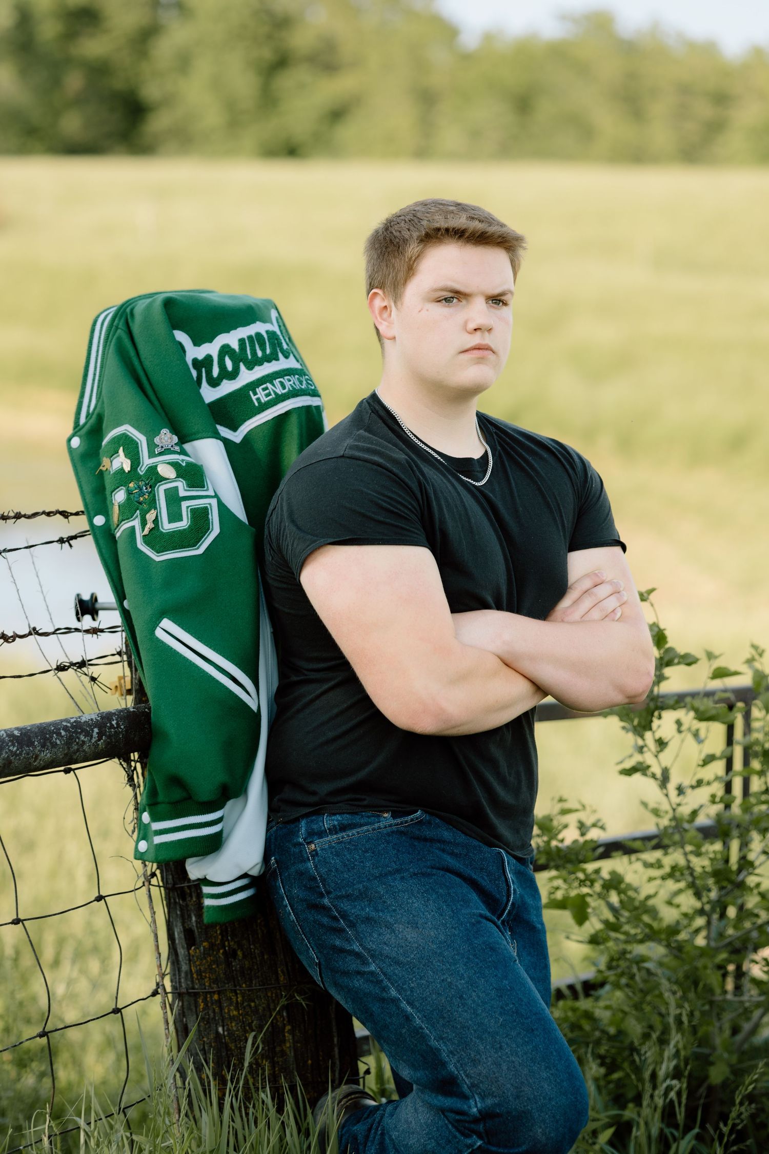 Serious teen boy in black shirt crosses arms leaning on fence with green letterman jacket beside him.