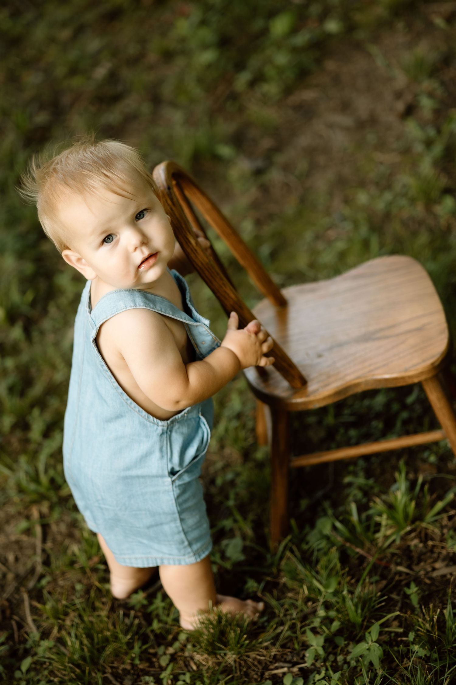 Toddler in blue denim romper standing barefoot on grass holding a wooden chair while looking up at camera.