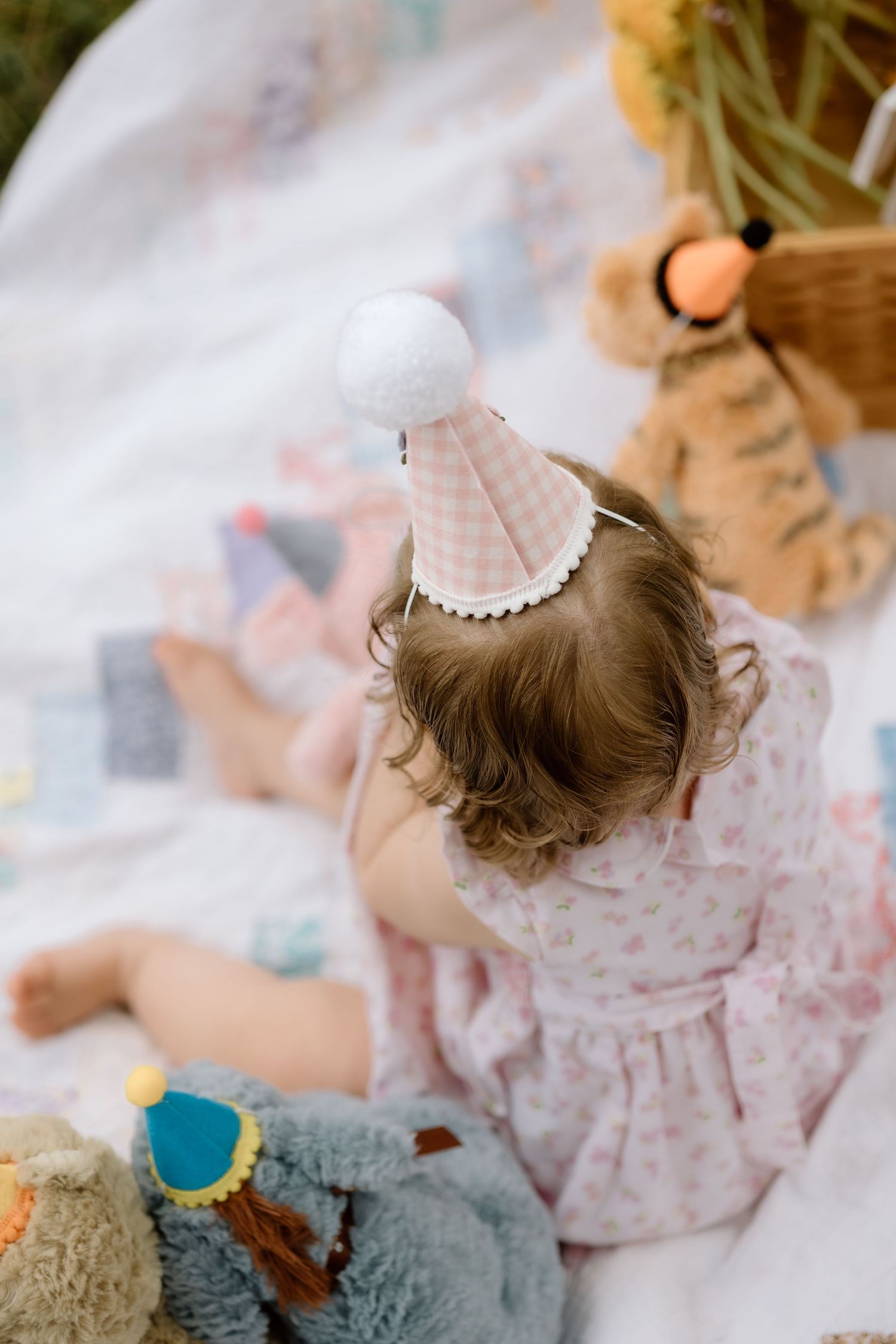 Overhead view of baby girl in pink gingham party hat sitting with toys on a quilt.
