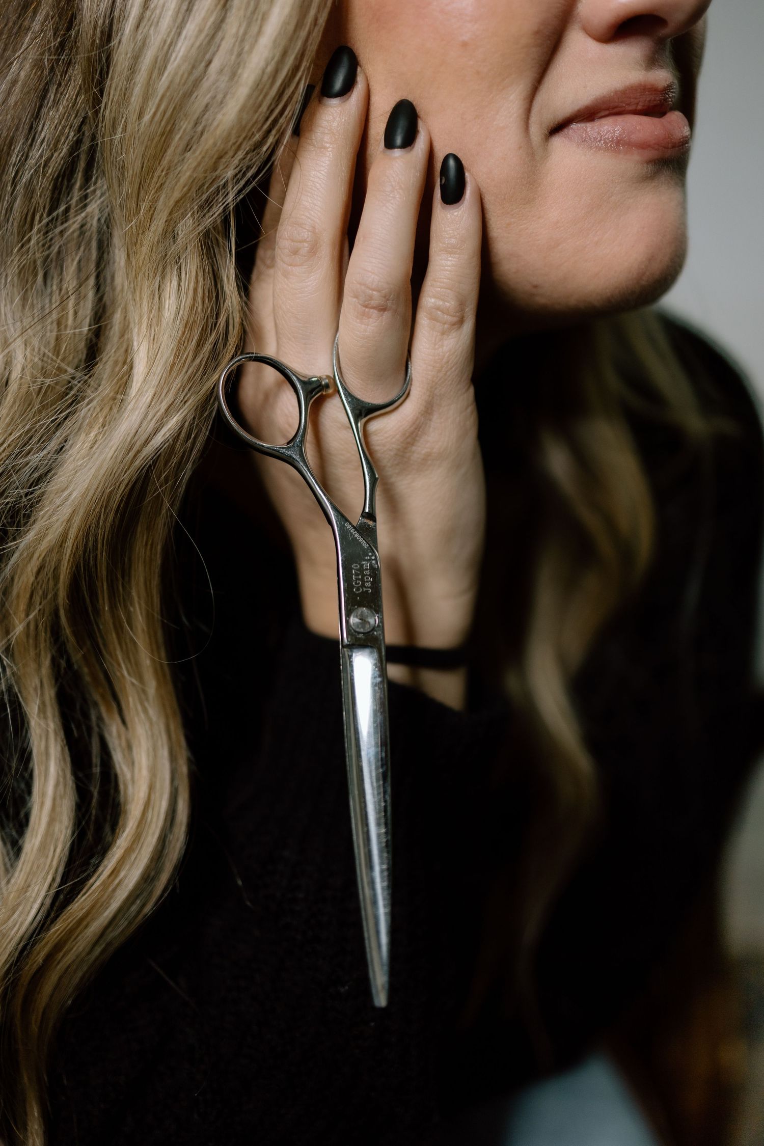 A woman with black nails holds professional hair scissors against her cheek, smiling softly against a light background.