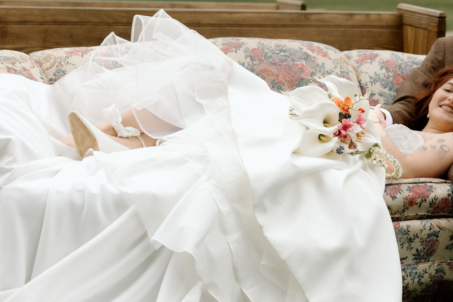 Bride in white wedding gown lying on floral sofa holding bouquet of calla lilies.