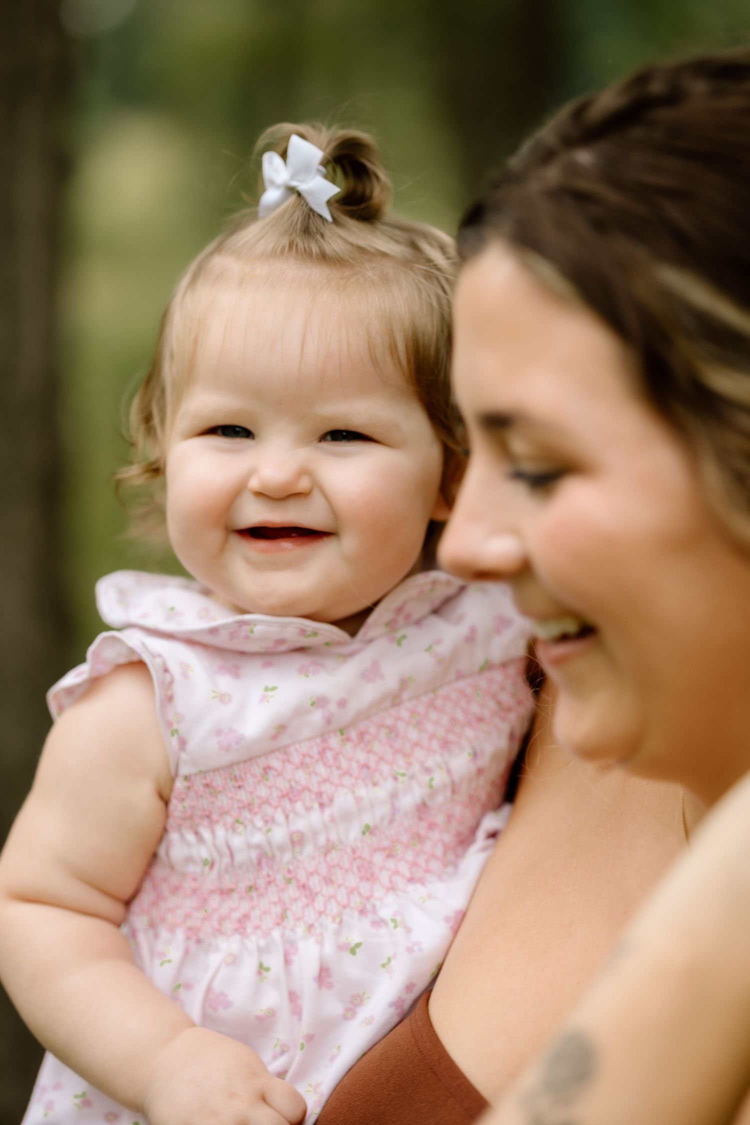 Smiling baby girl with white bow held by her mother outdoors in a pink smocked dress.