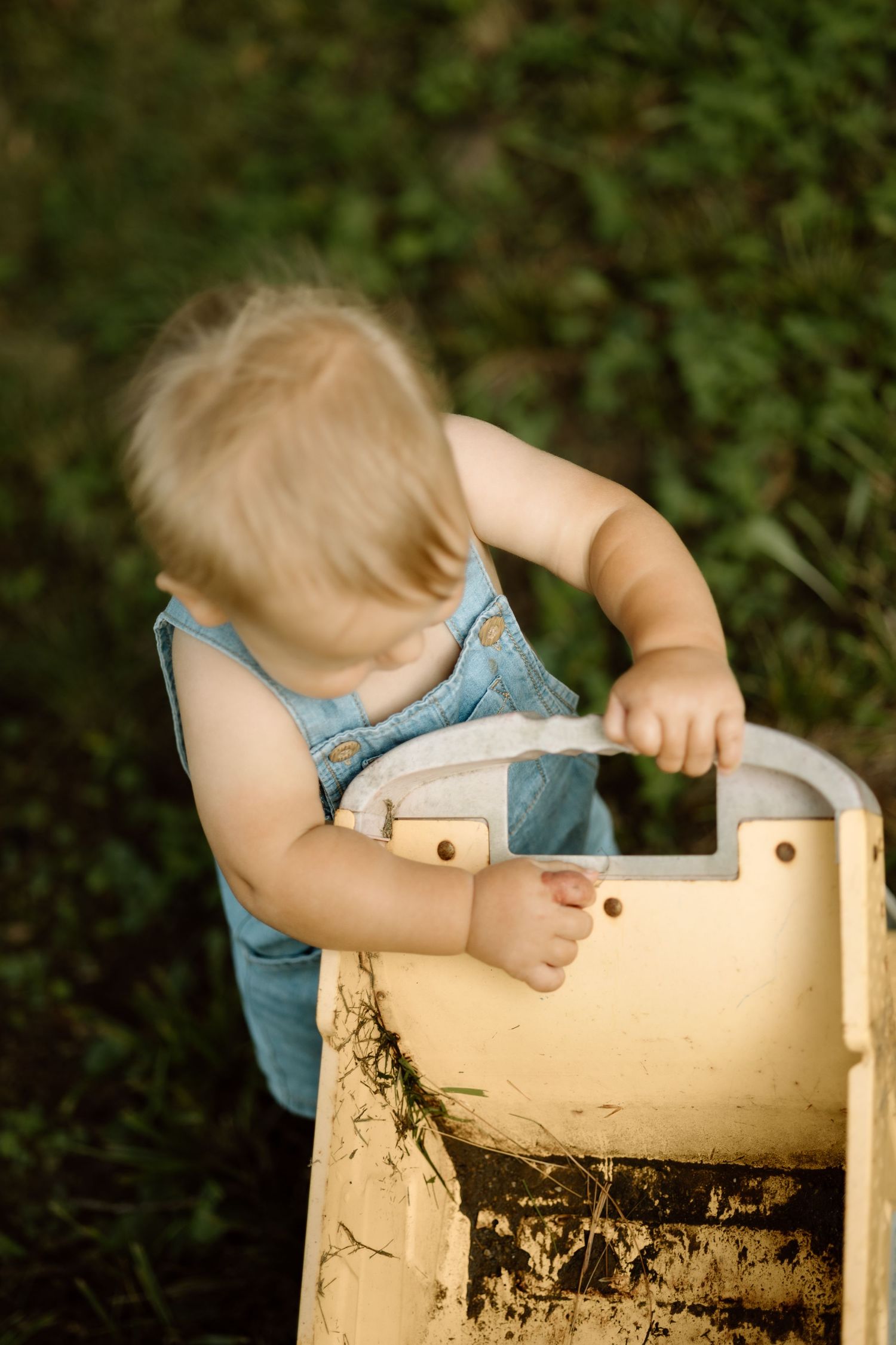 Toddler in blue denim romper leaning over and exploring a yellow wooden toy outdoors on green grass.