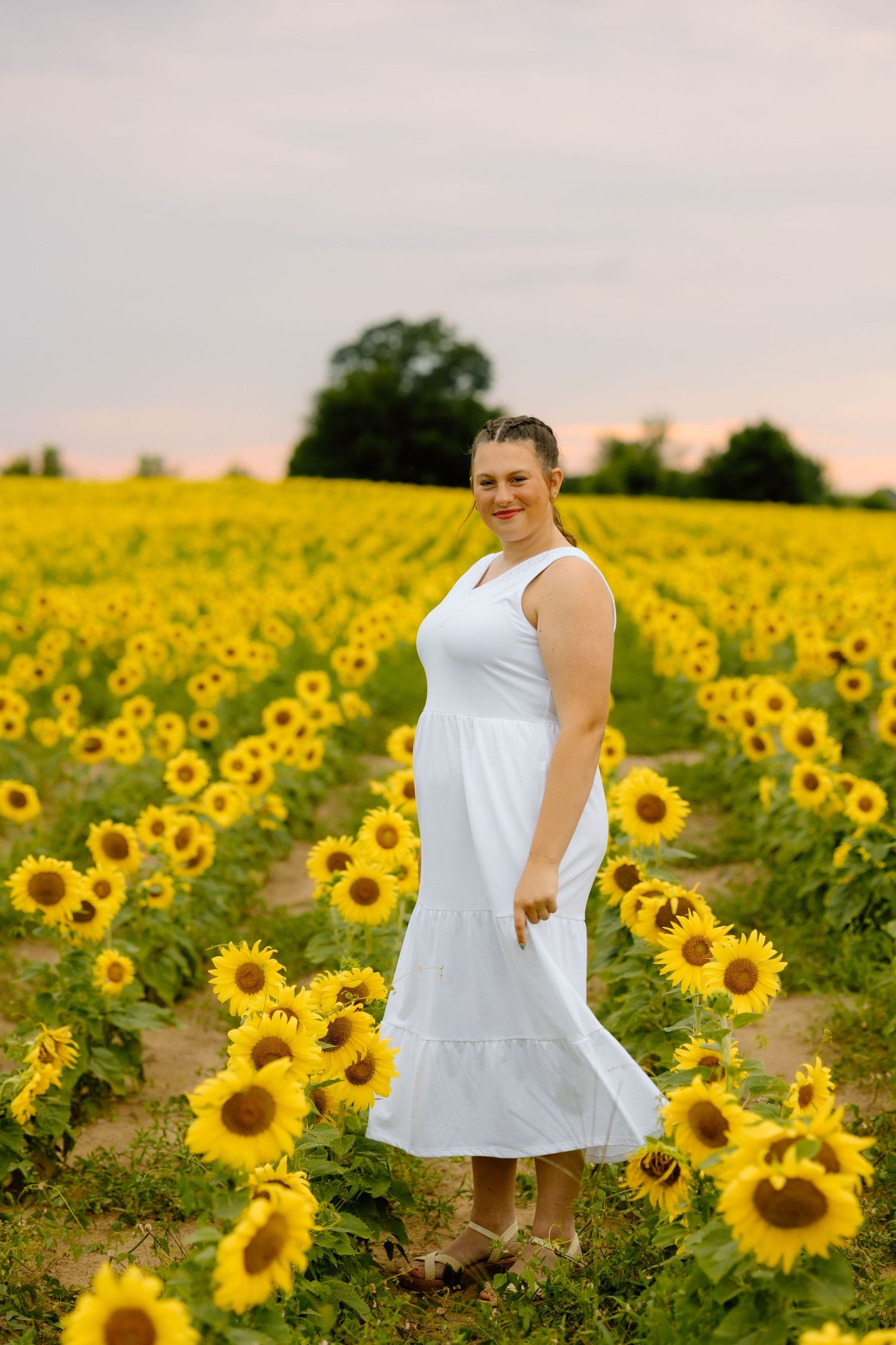 Smiling woman in white dress standing in vibrant yellow sunflower field at sunset.