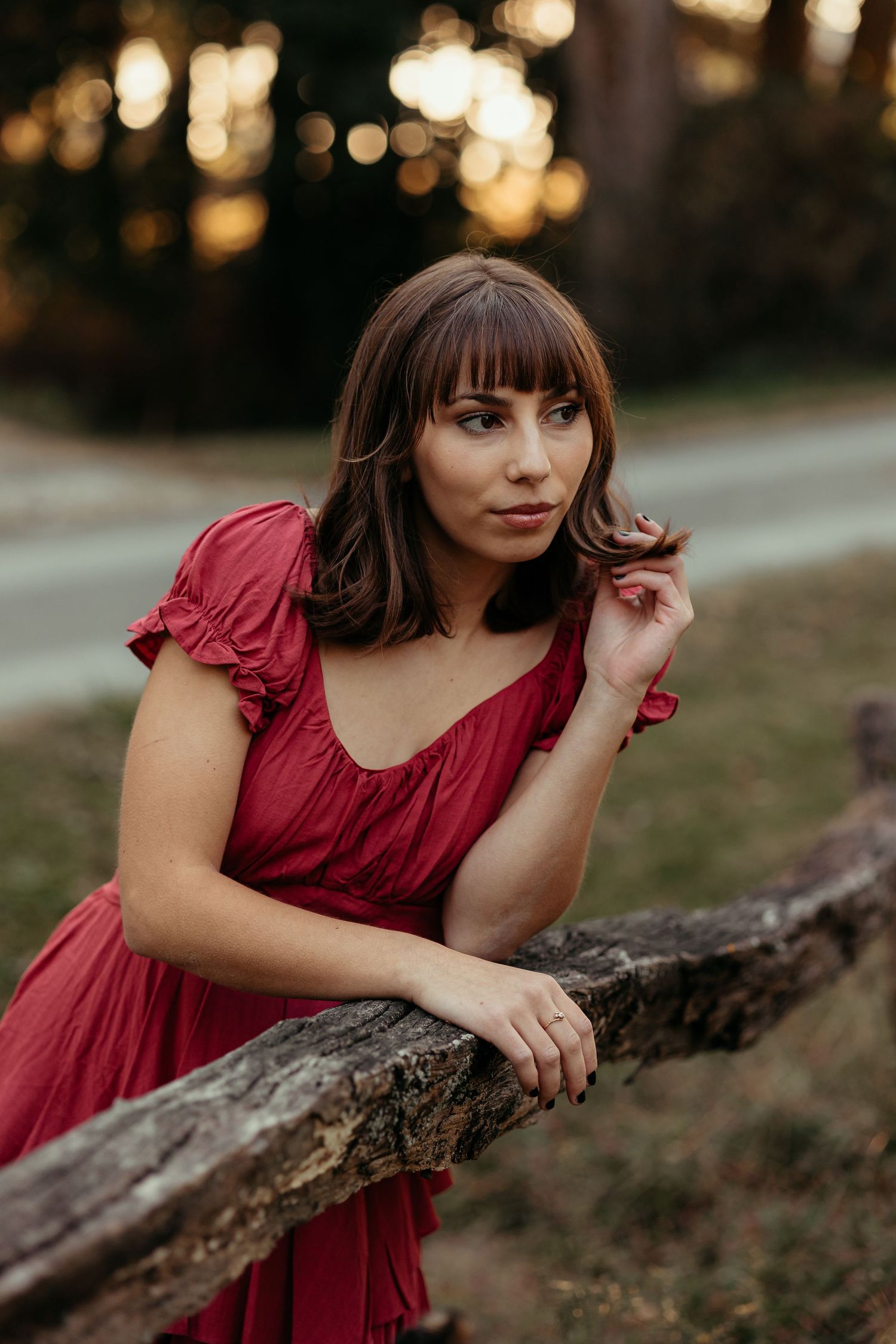 Young woman in red dress leaning on a wooden fence touching her hair at dusk.