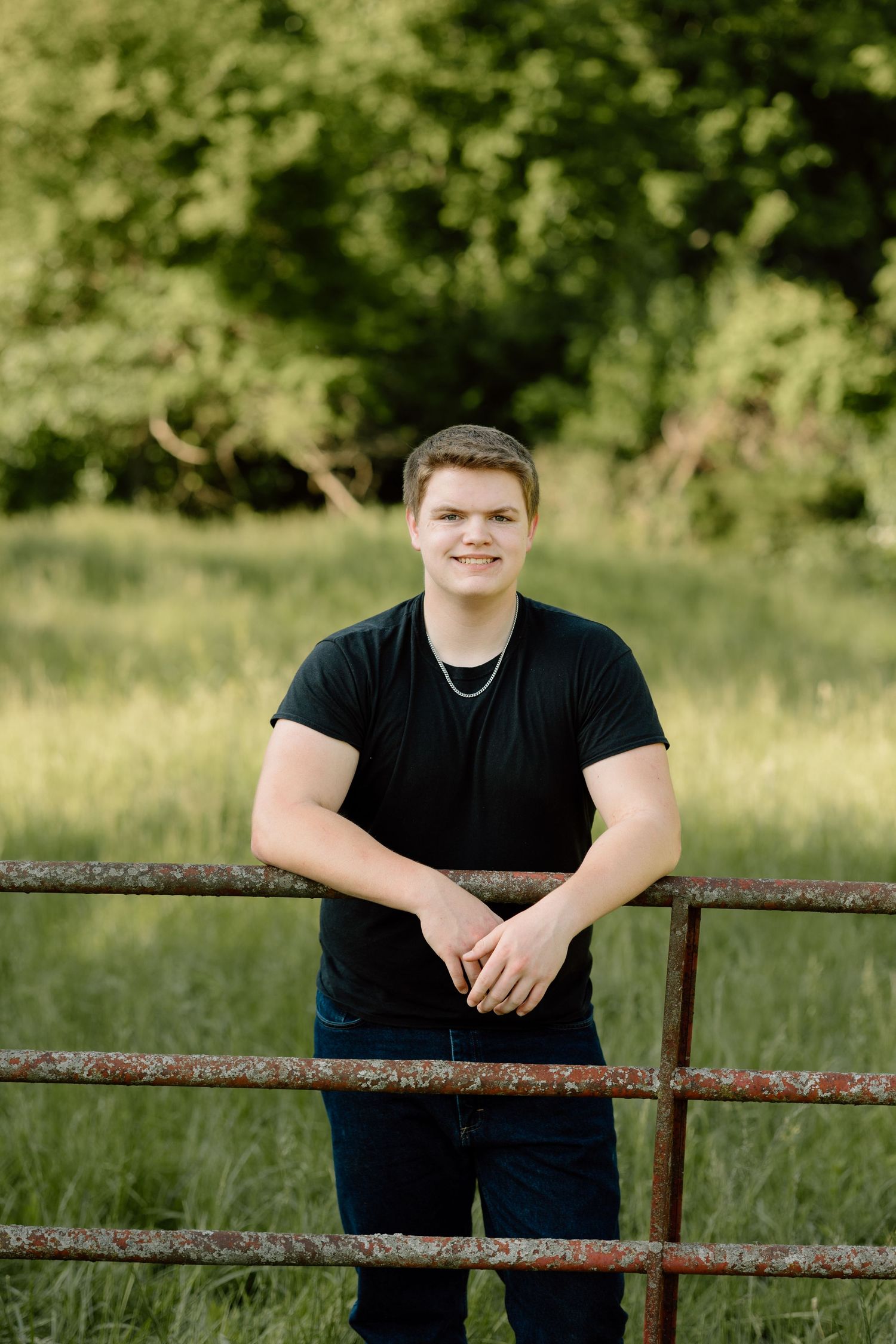 Smiling young man in black t-shirt leans on rusty metal gate in lush green field.