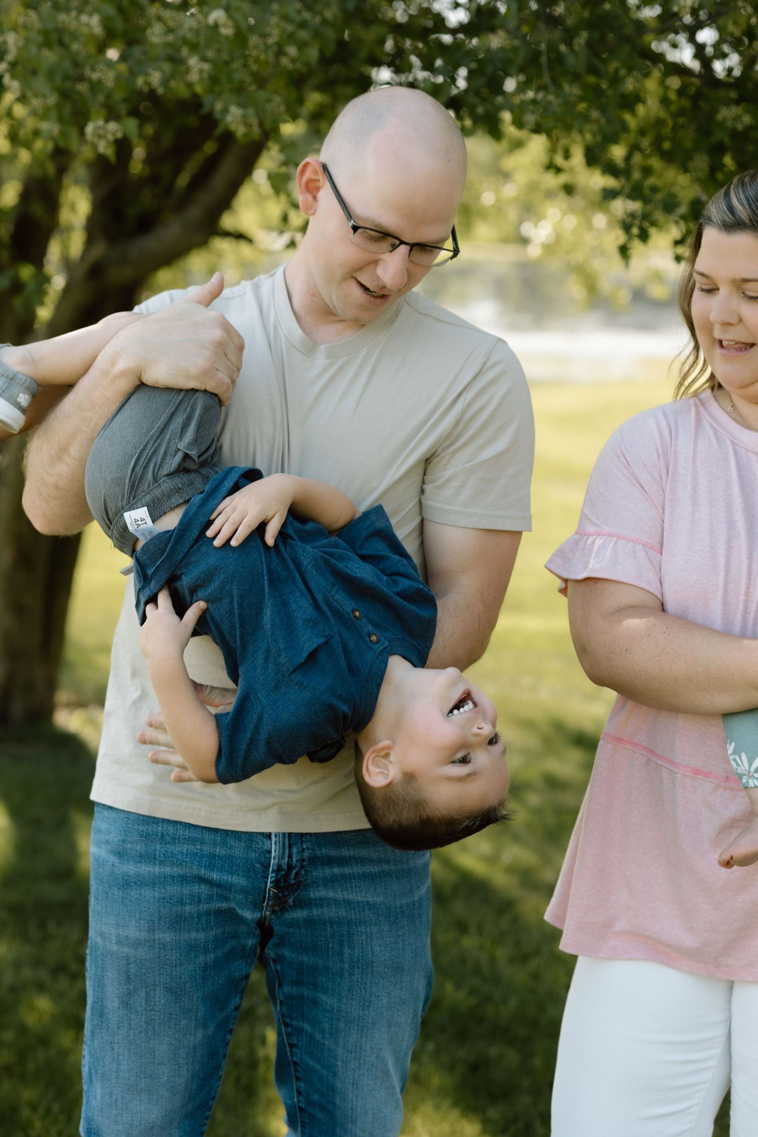 Laughing father playfully holds young son upside down while mother watches and smiles in sunny park.