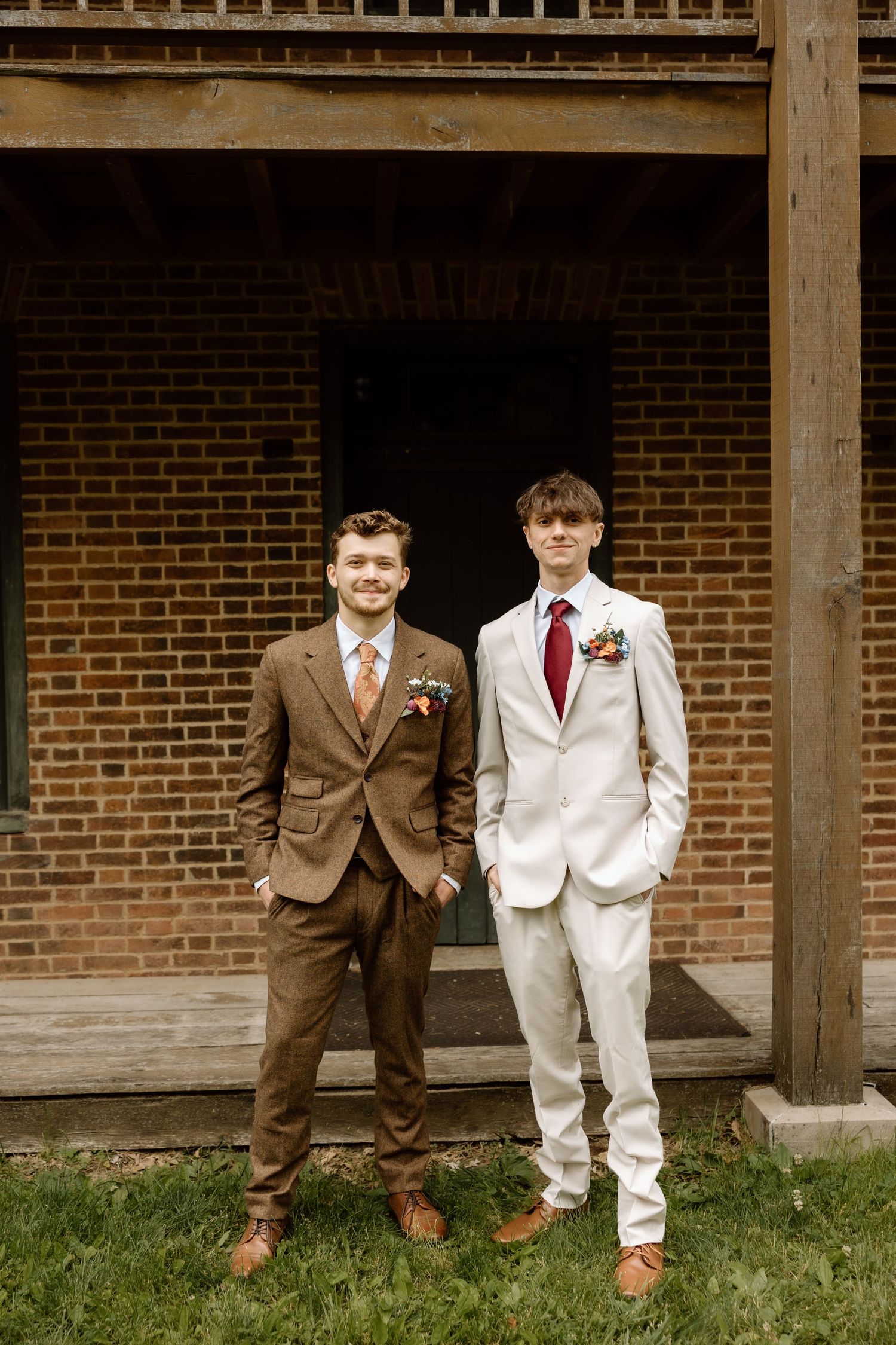 Two groomsmen posing before brick building, one in brown tweed suit and one in ivory suit with red tie.