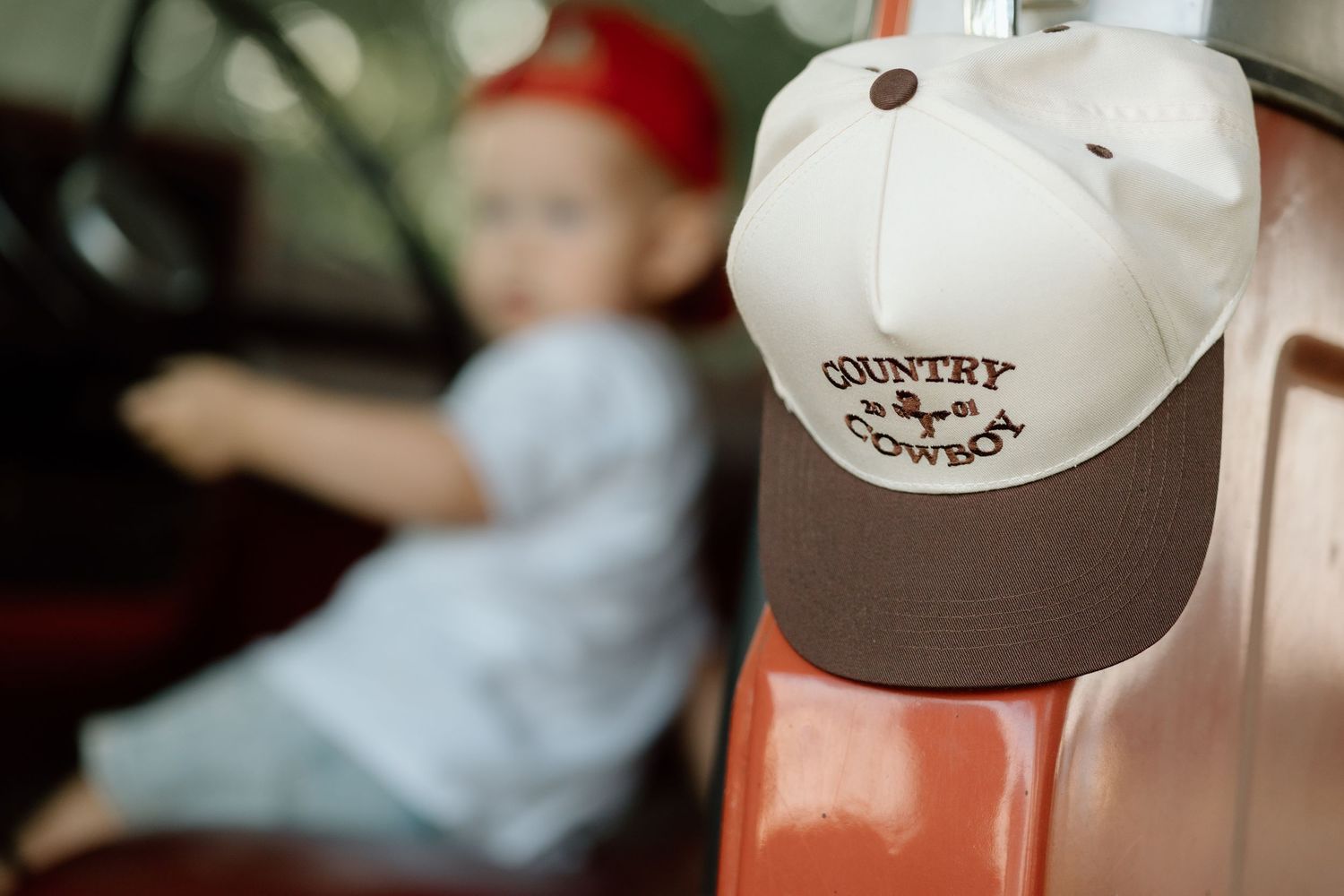 Country Cowboy cap hangs on red truck door with toddler sitting inside in background.