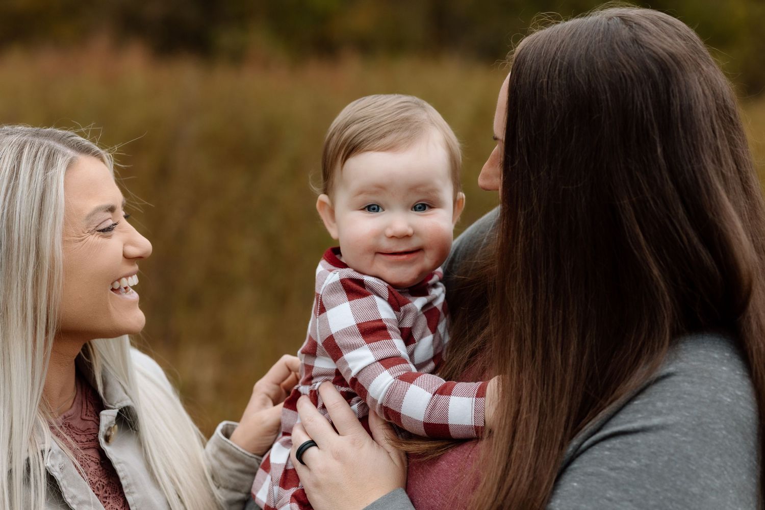 Two moms smile lovingly at their cute baby during a golden autumn family photography session.