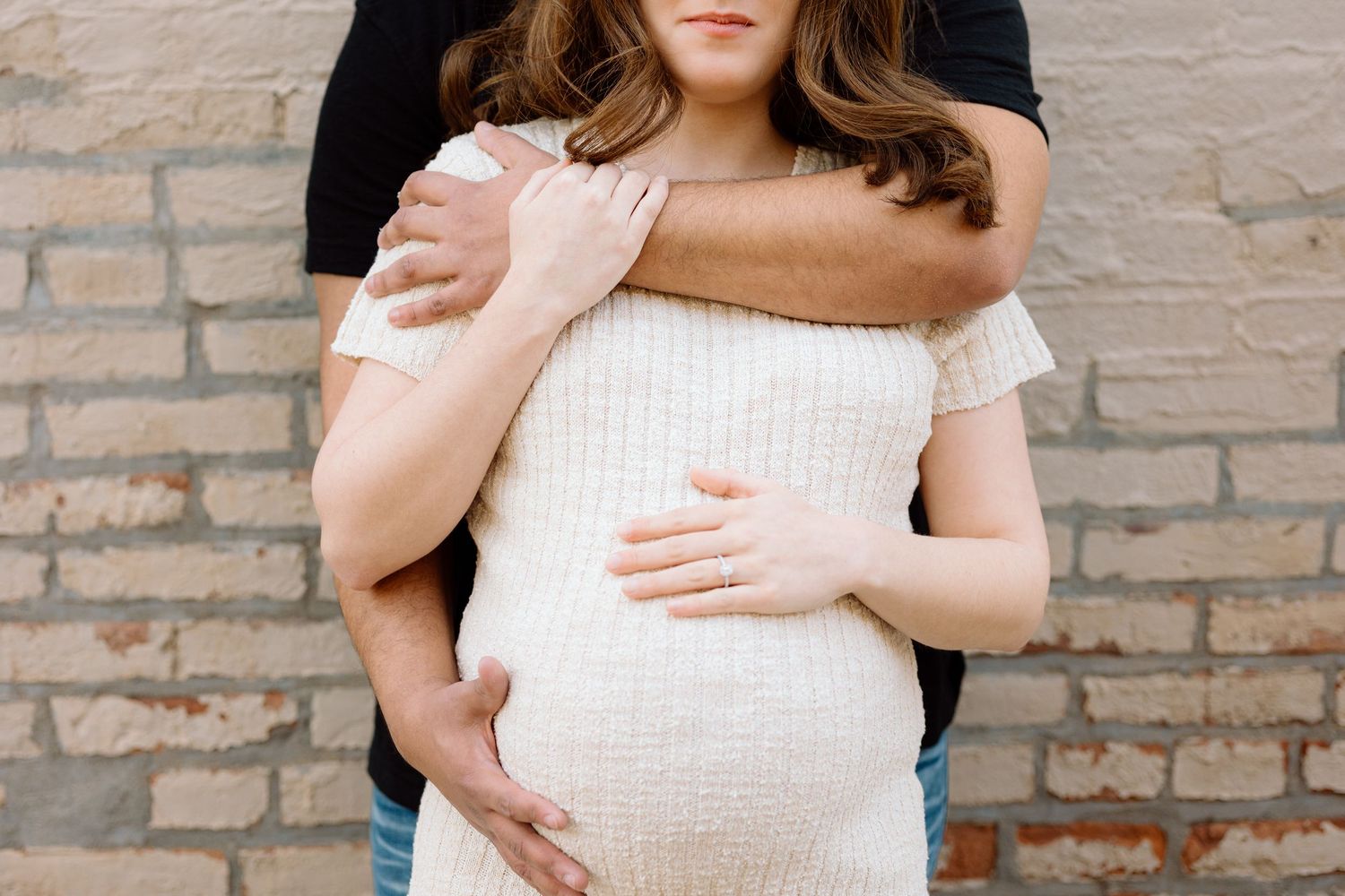 Close-up of pregnant woman in white textured dress with partner embracing her from behind near brick wall.