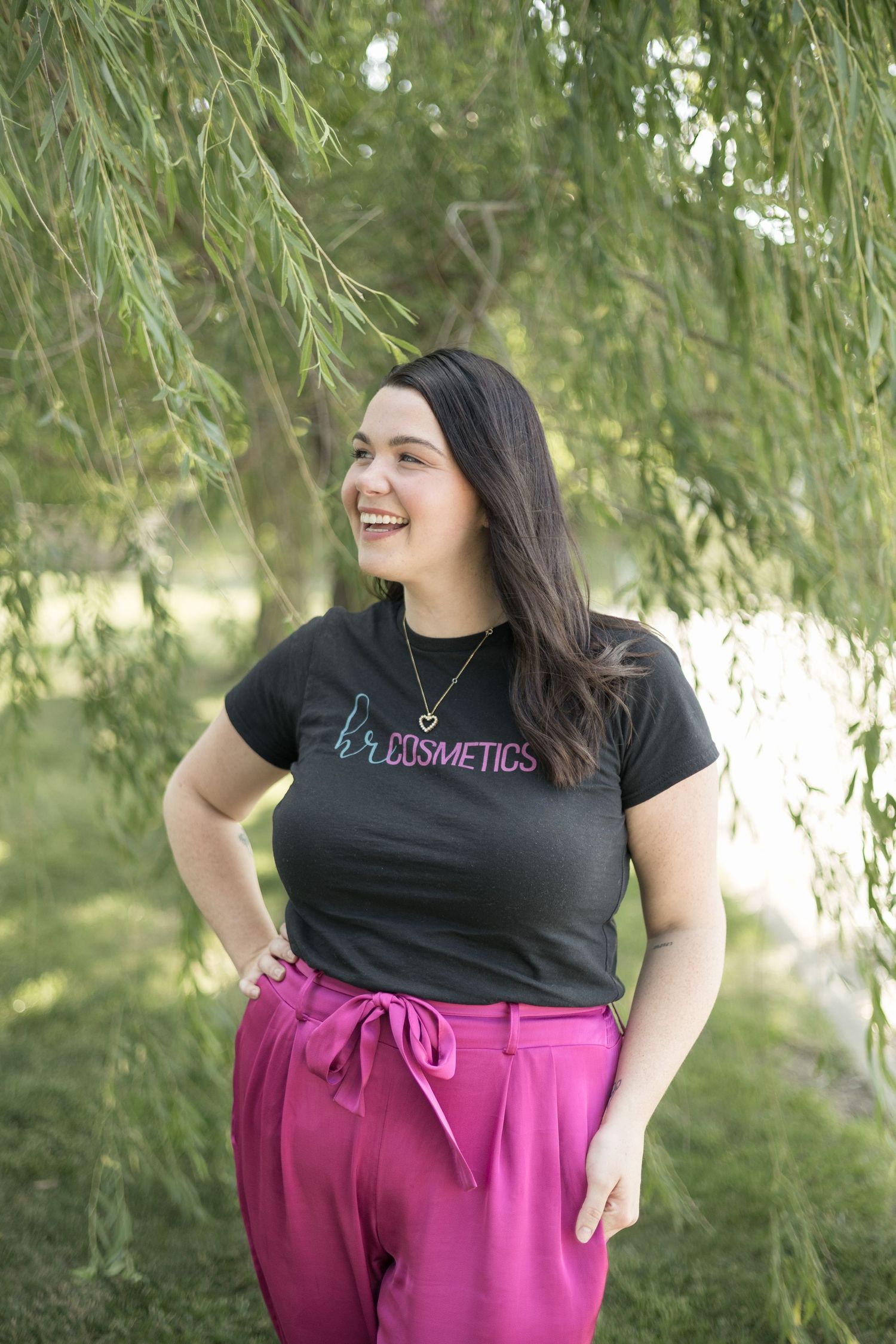 Laughing woman in HR Cosmetics shirt and pink pants standing under weeping willow branches outdoors.
