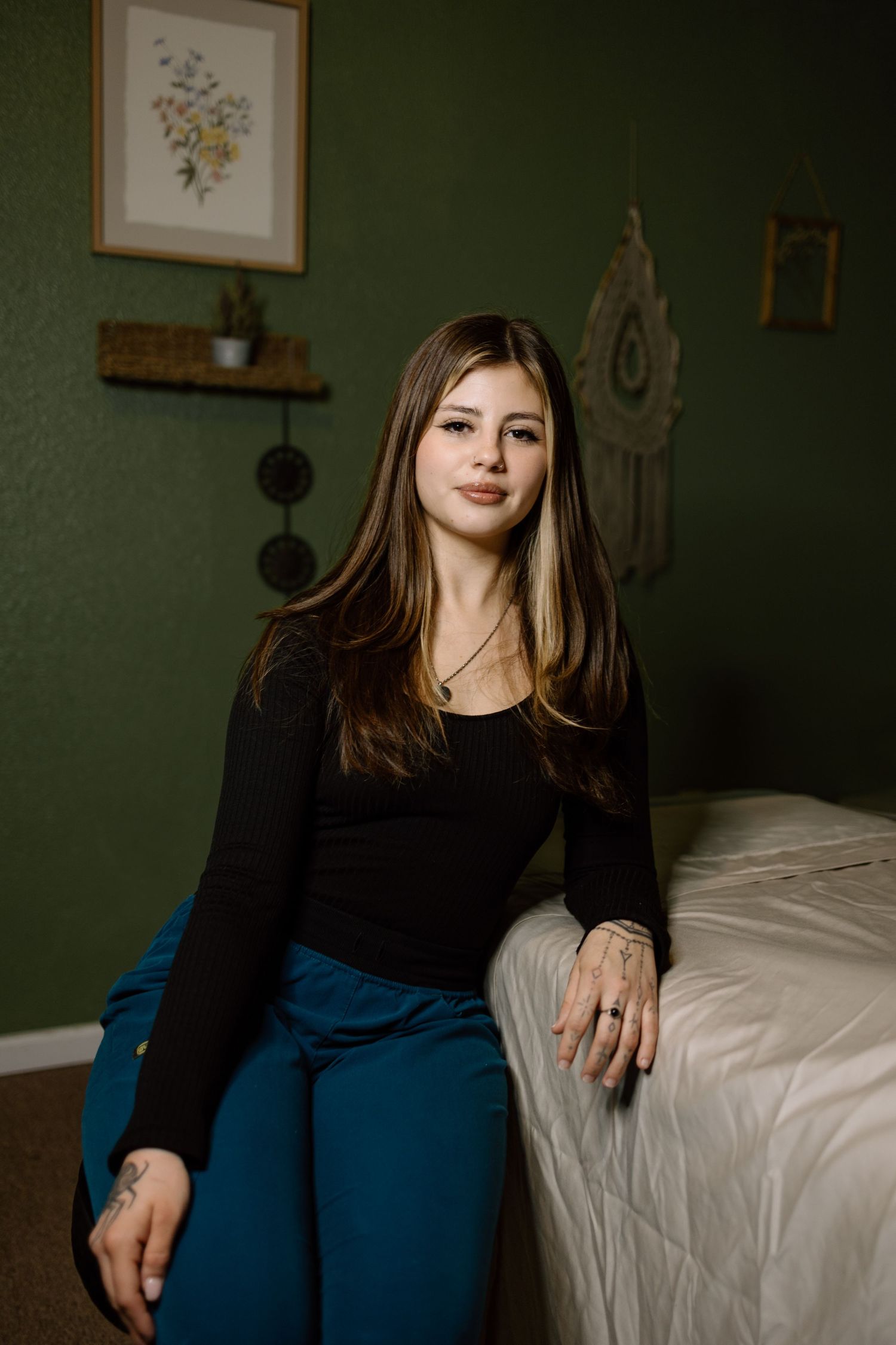 Young woman with highlighted hair wearing black top and teal scrub pants sitting beside massage table.