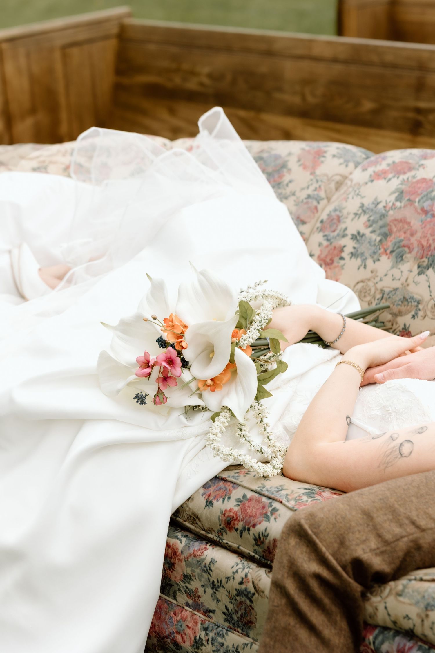 Bride holding a white and orange wedding bouquet while resting on a vintage floral sofa in a white gown.