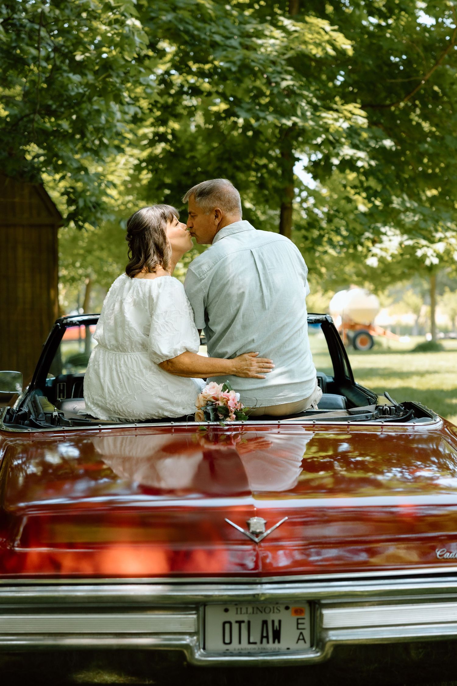 Newlywed couple kissing while seated on trunk of red classic Cadillac convertible with OTLAW plate.