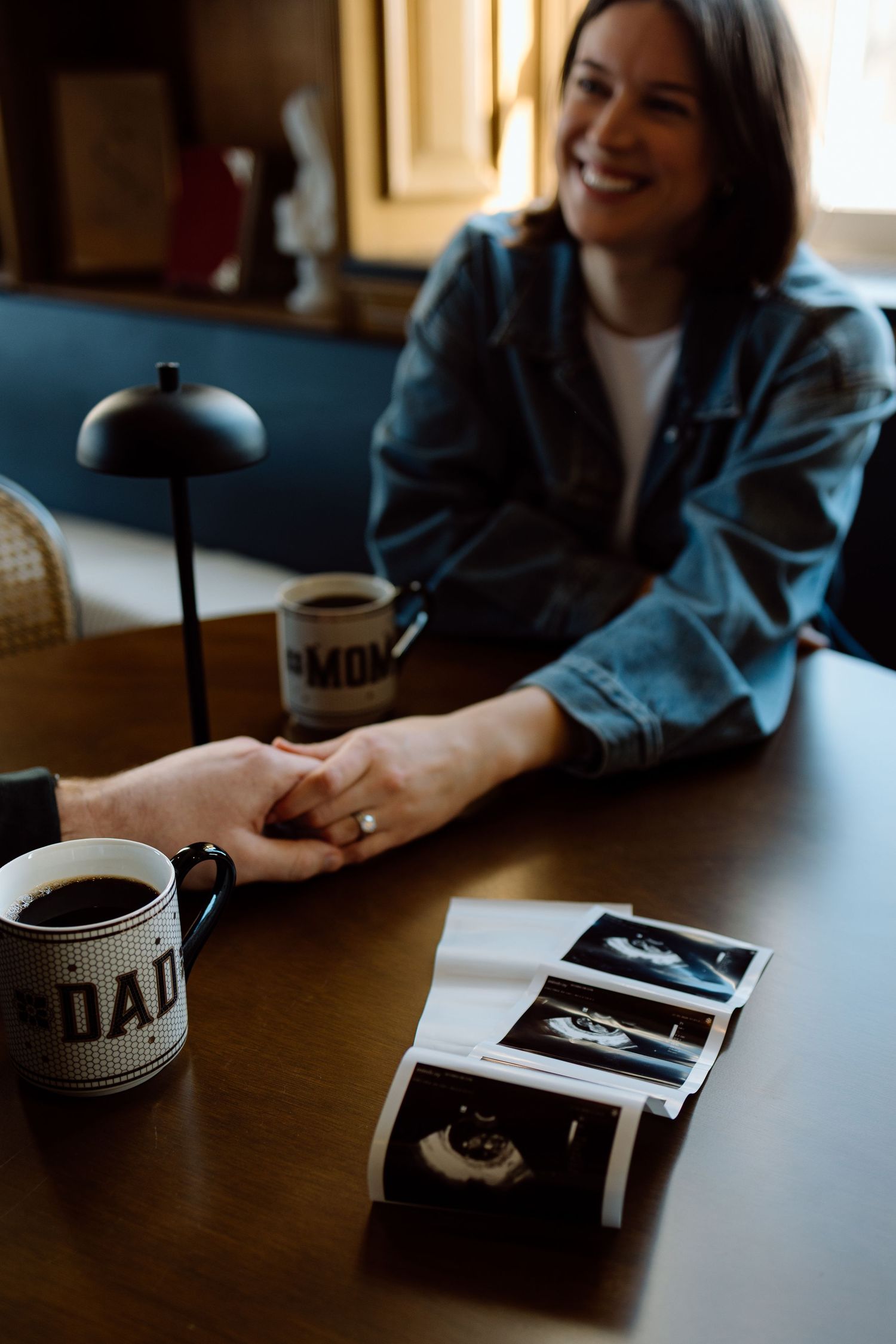Smiling woman in denim jacket holds partner's hand at table with Mom mug and ultrasound photos.