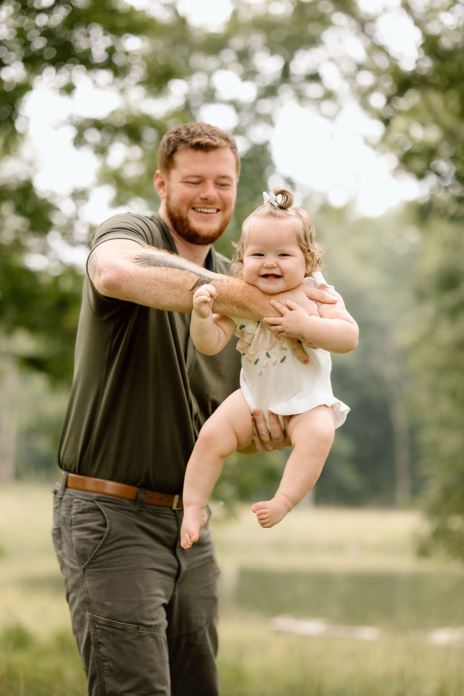 Smiling father holds happy baby girl up in a sunny green outdoor park setting.