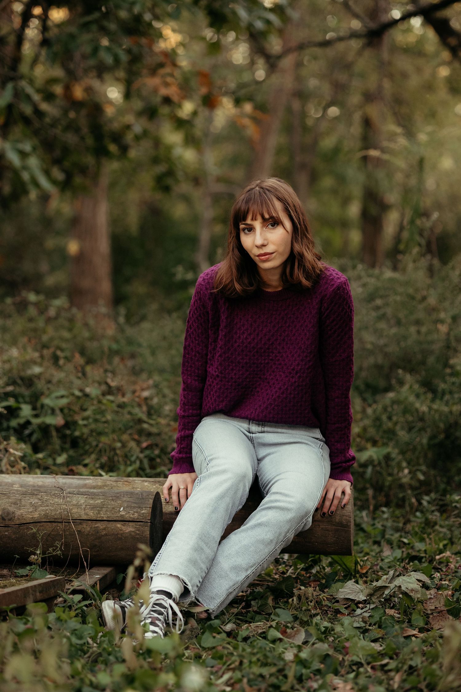Young woman in purple sweater and jeans sitting on a log in the forest.