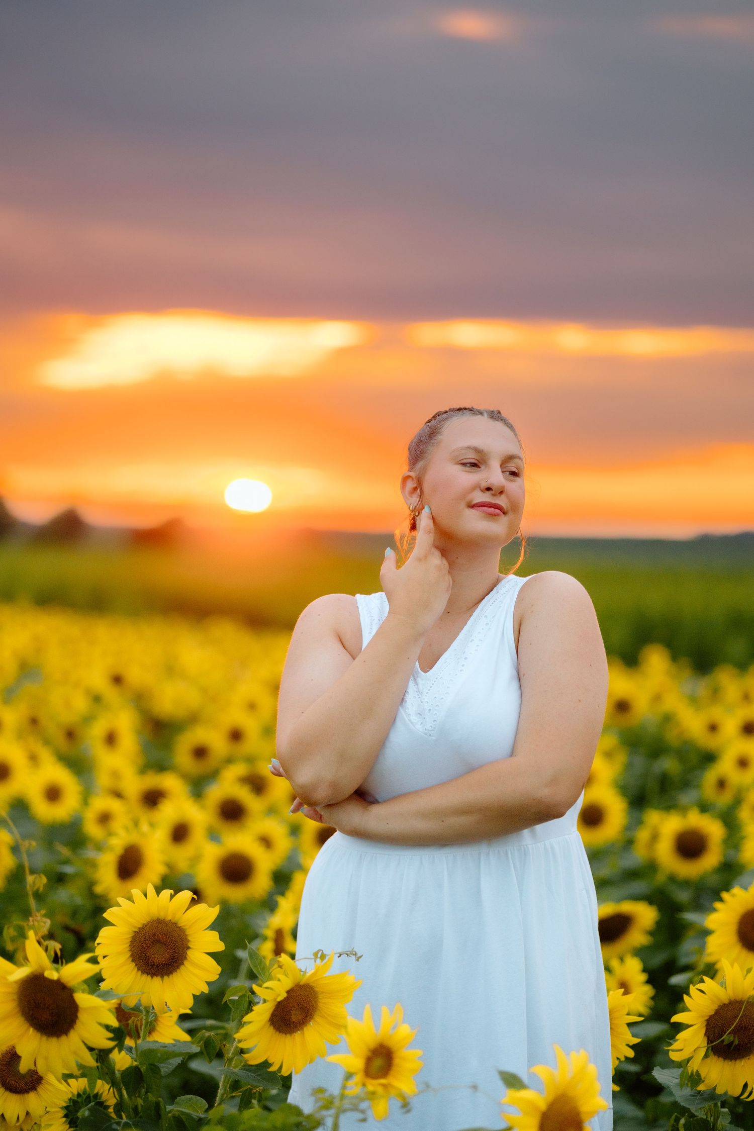 Serene woman in white dress with eyes closed standing in a sunflower field at sunset.