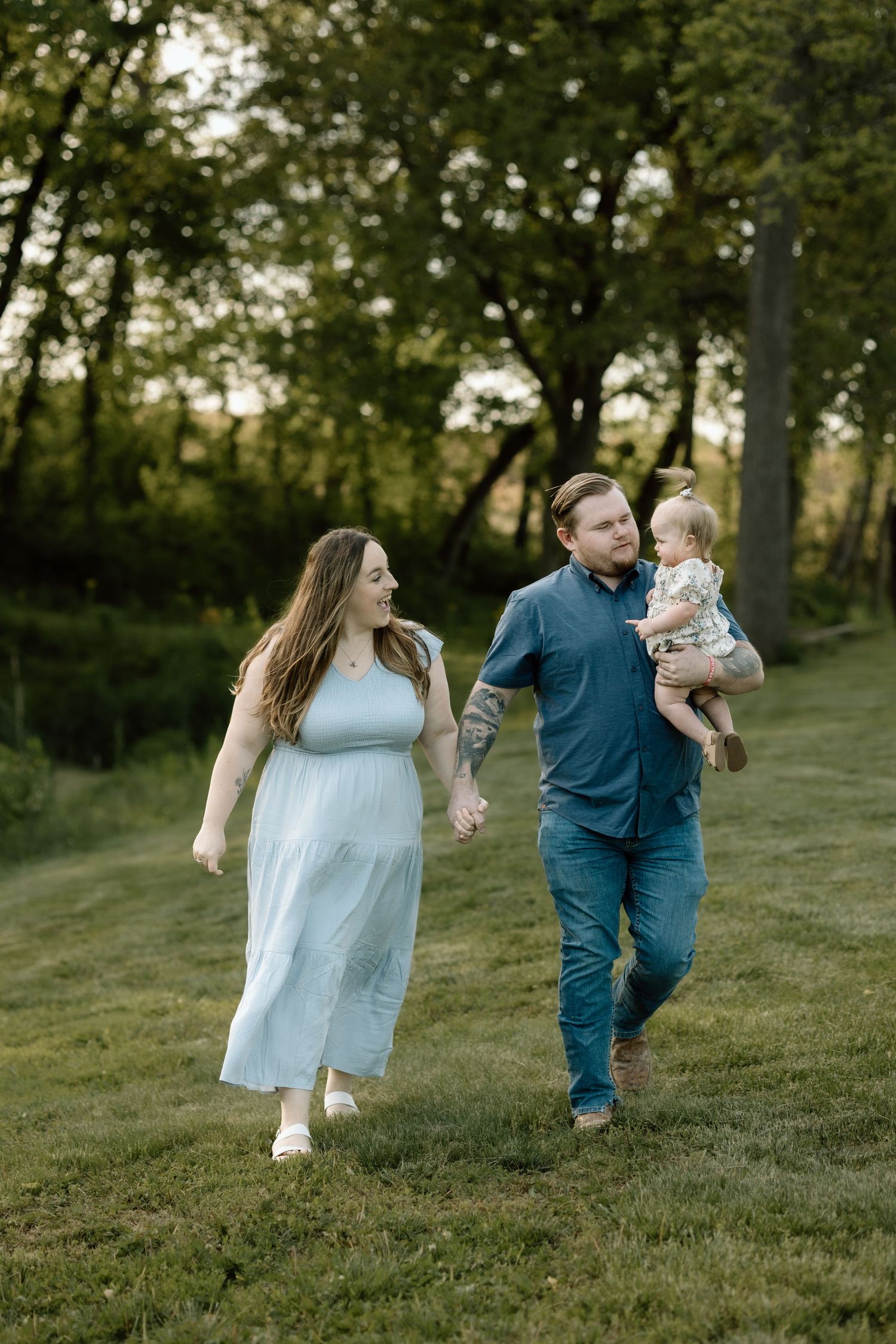 A happy family walks hand-in-hand through a sunlit green field, father carrying their baby girl at golden hour.