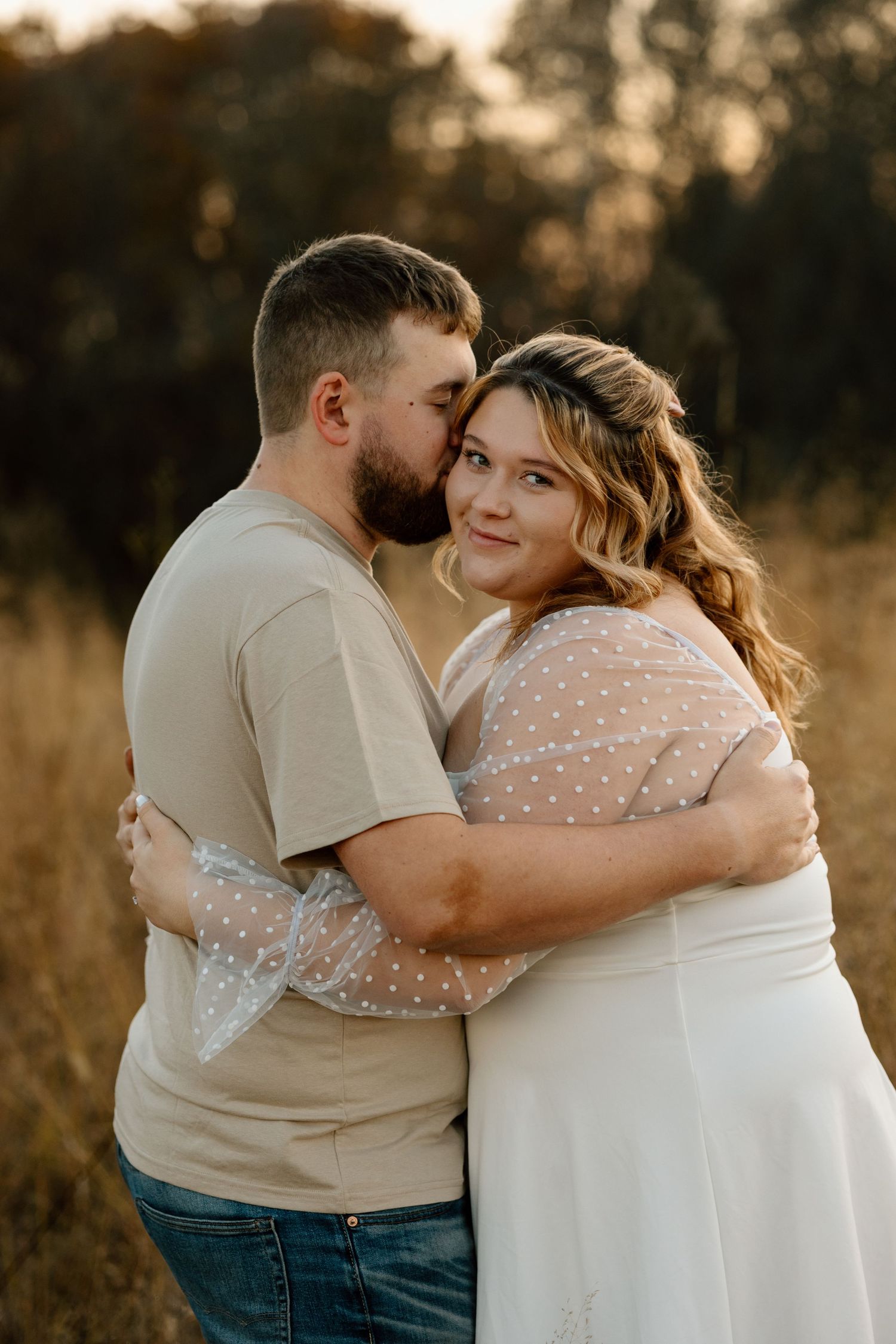 Bearded man kissing woman on cheek as she smiles at camera during golden hour engagement photo session outdoors.