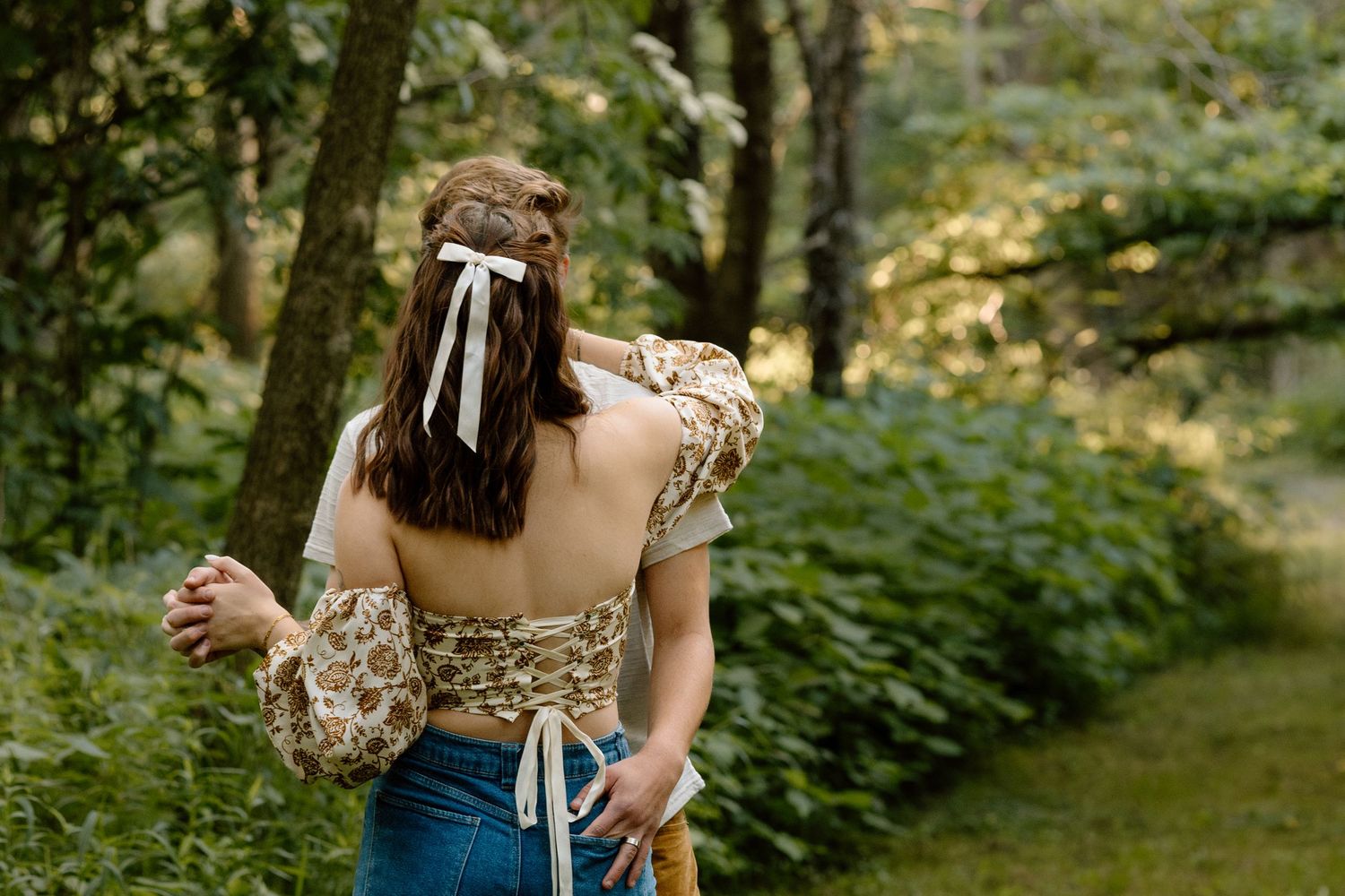 Woman with white ribbon hair bow in floral crop top walking through green forest path.