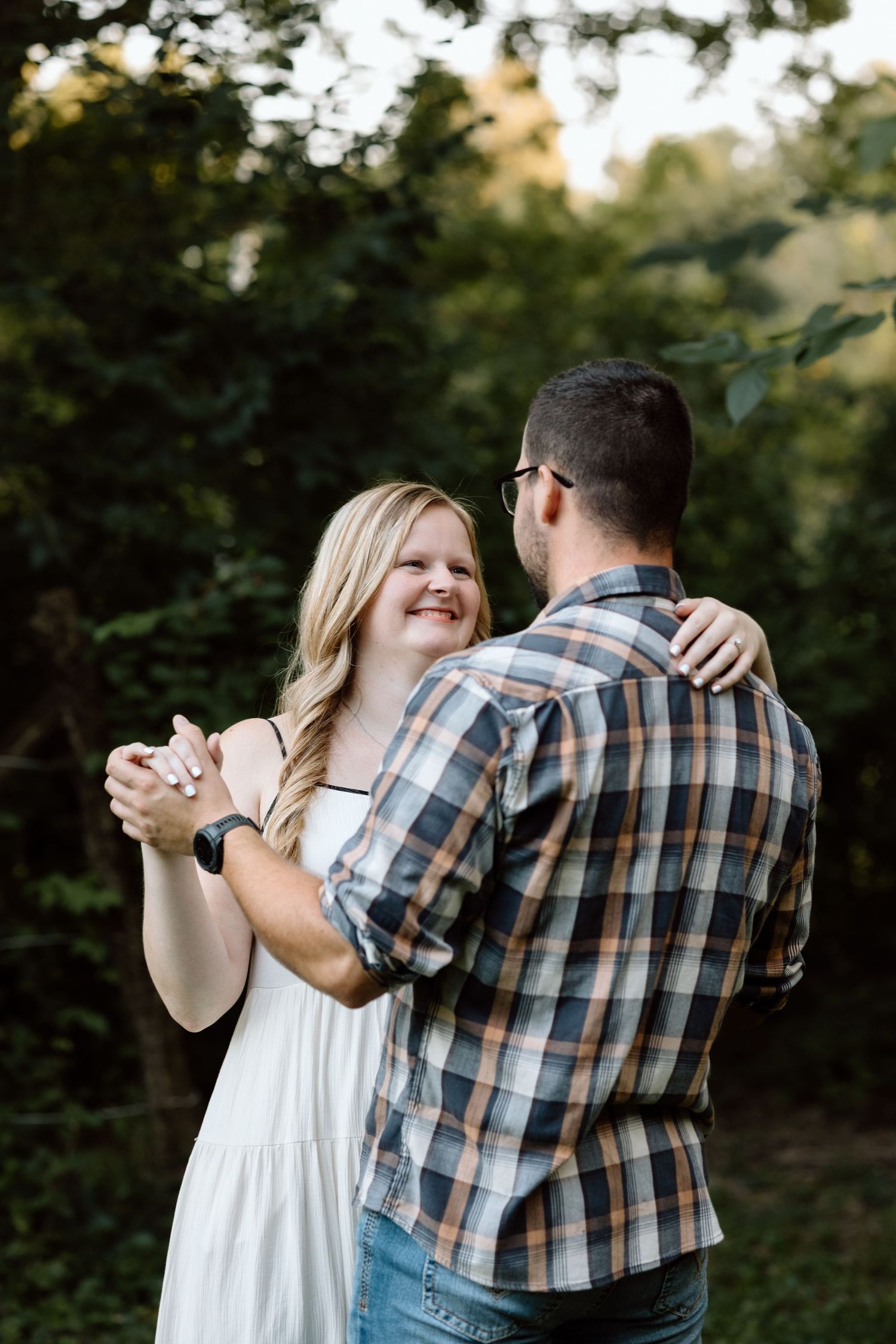 A smiling couple dances outdoors, the woman in a white dress holding hands with a man in a plaid shirt, surrounded by lush greenery.