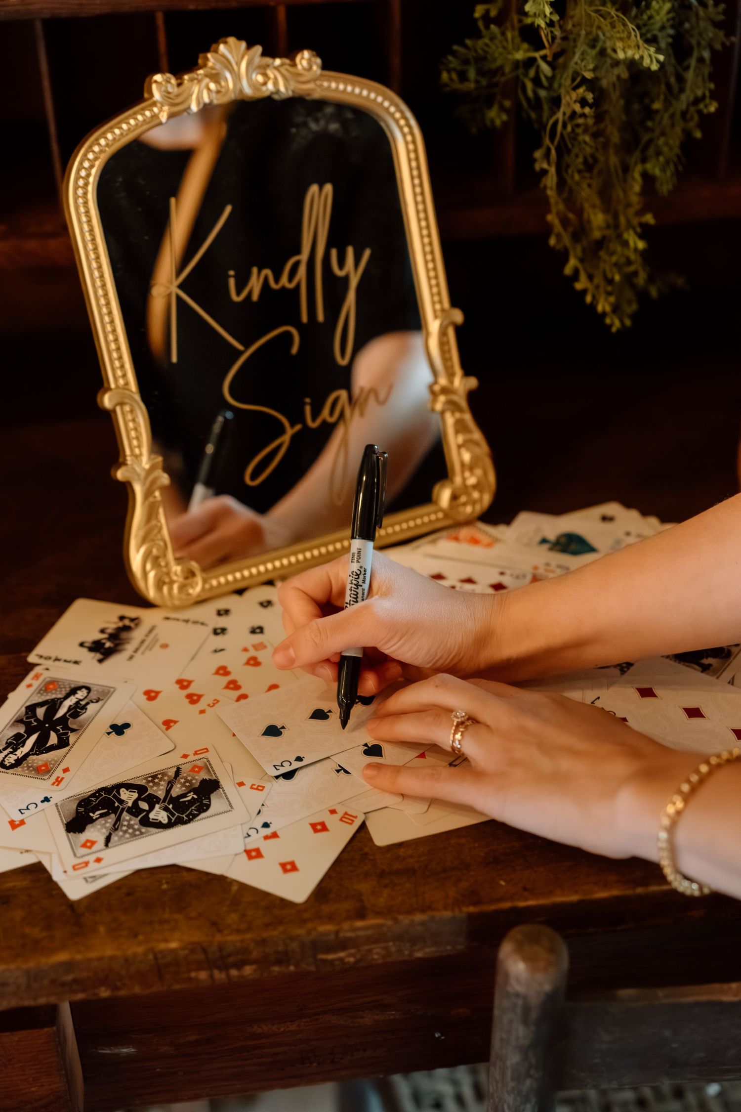 Wedding guest signing playing card guestbook beside ornate gold mirror with Kindly Sign inscription.