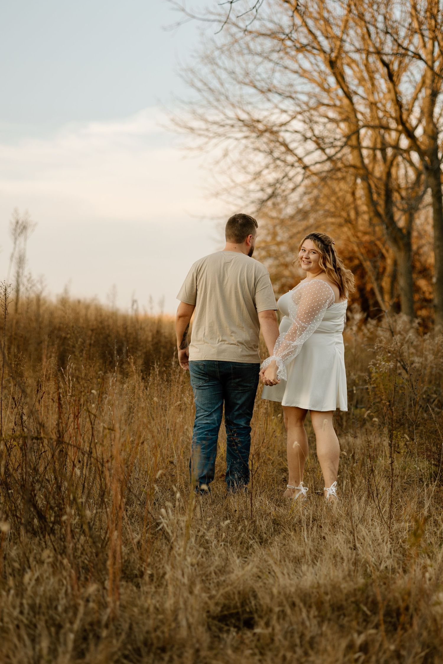Couple holding hands walking through golden autumn field at sunset, woman in white dress smiling back at camera.