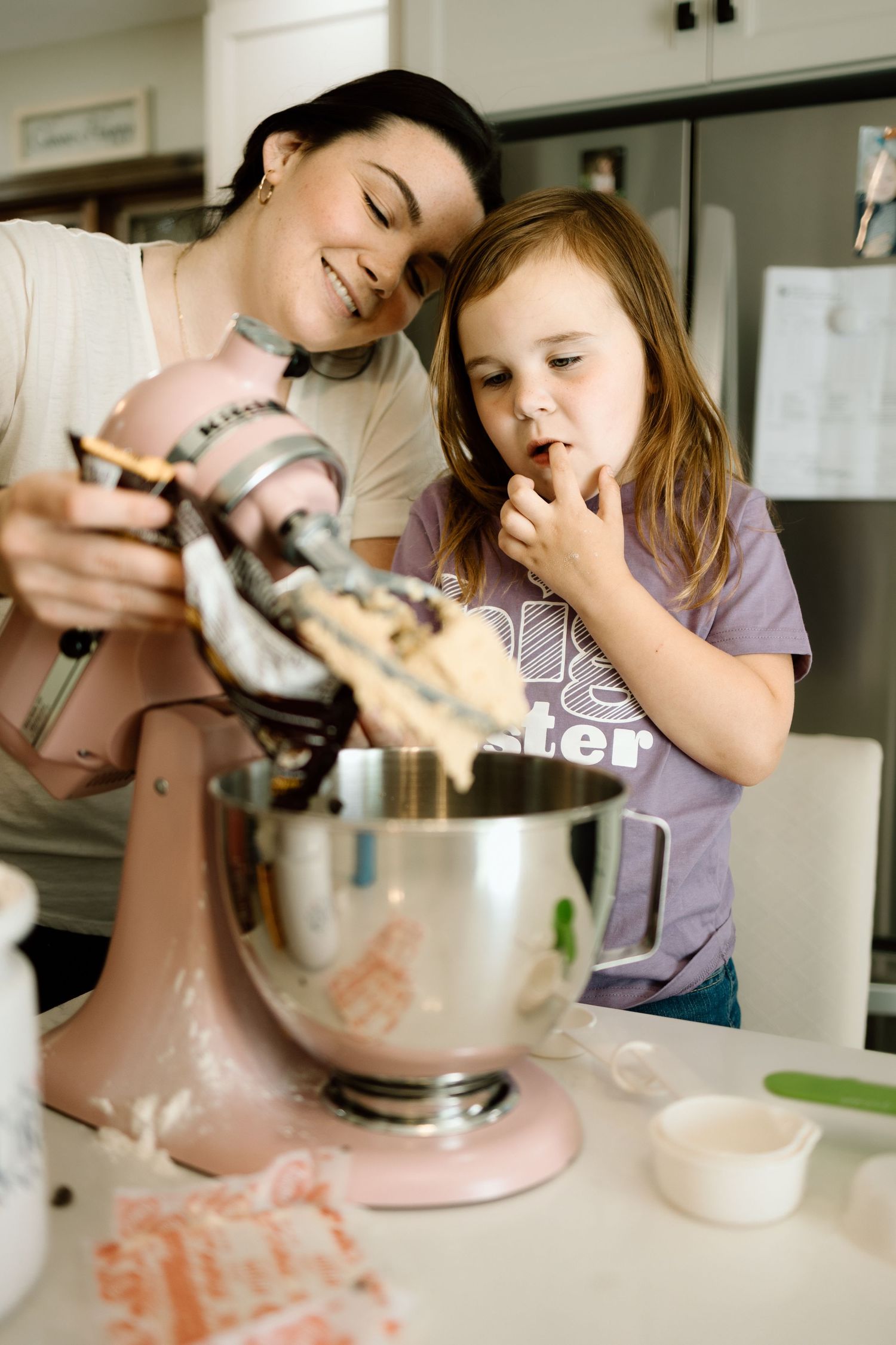 Smiling mother and young daughter tasting cookie dough from a pink KitchenAid stand mixer in kitchen.