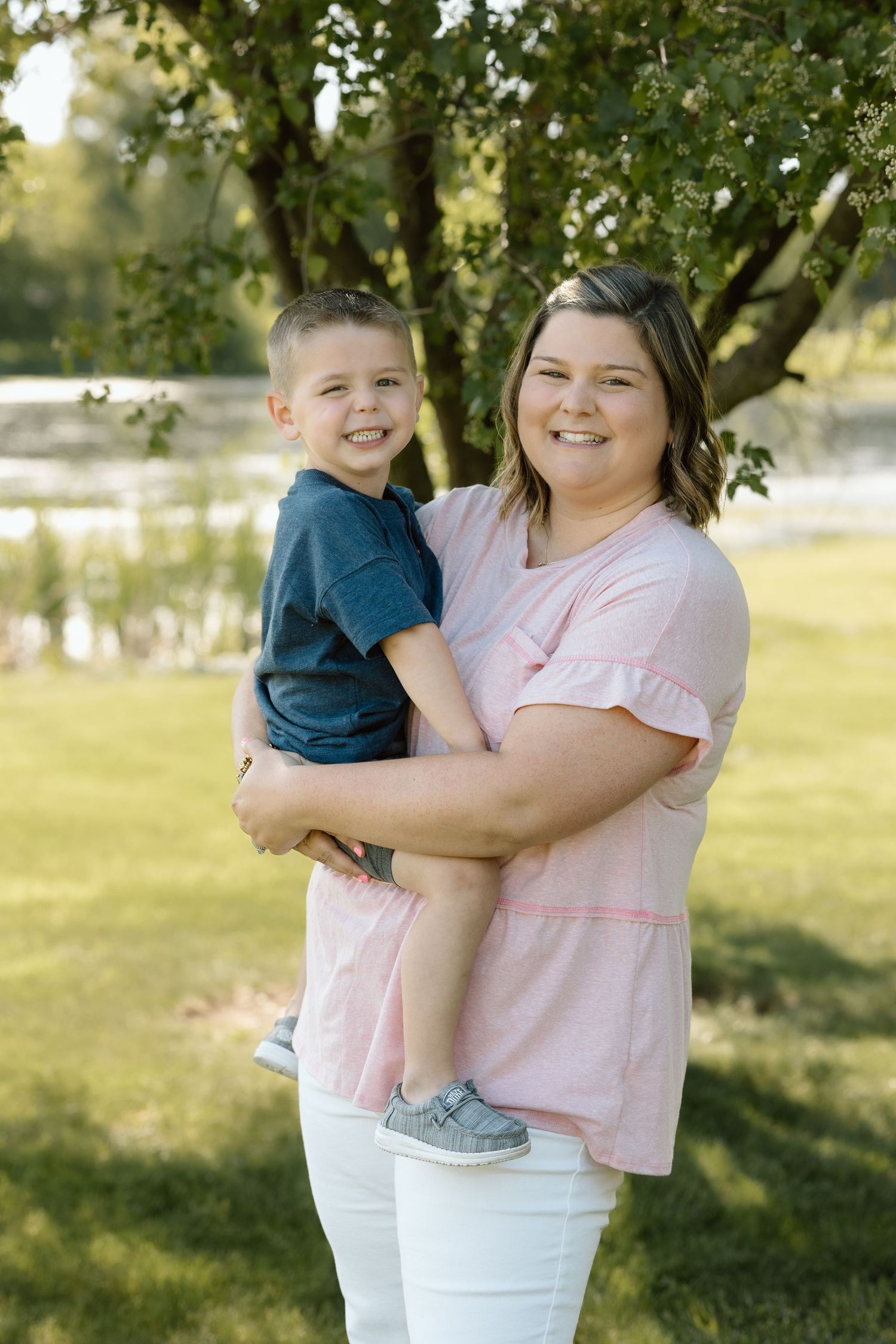 Smiling mom holds her young son in a navy shirt during a sunny outdoor family portrait session by a river.
