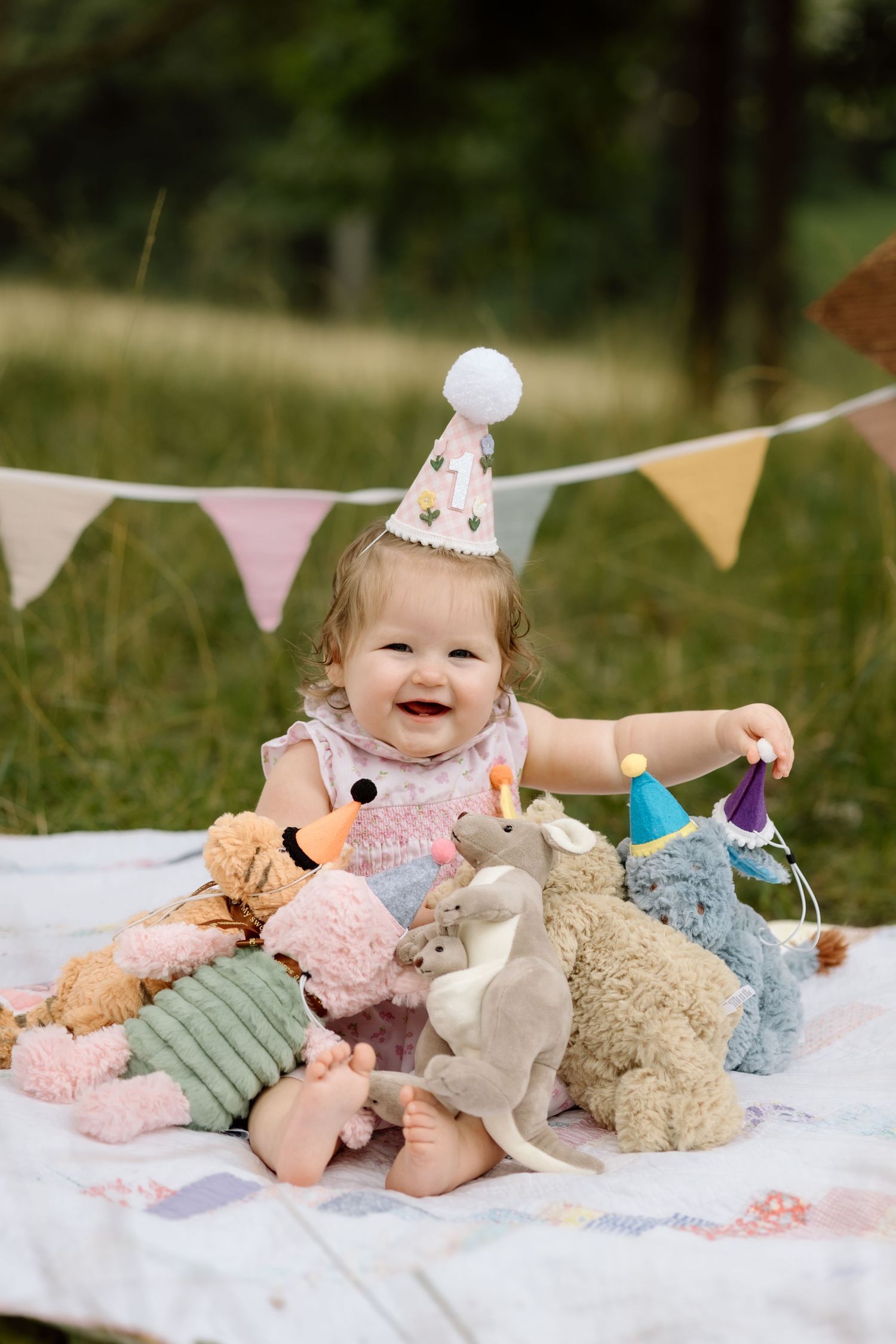 Smiling baby girl in party hat sits with stuffed animals on a blanket at outdoor birthday.