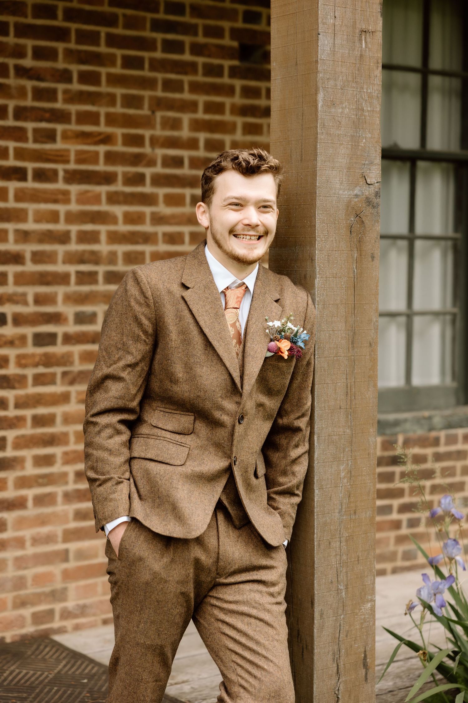 Smiling groom in brown herringbone tweed suit with colorful boutonniere leaning against wooden column.