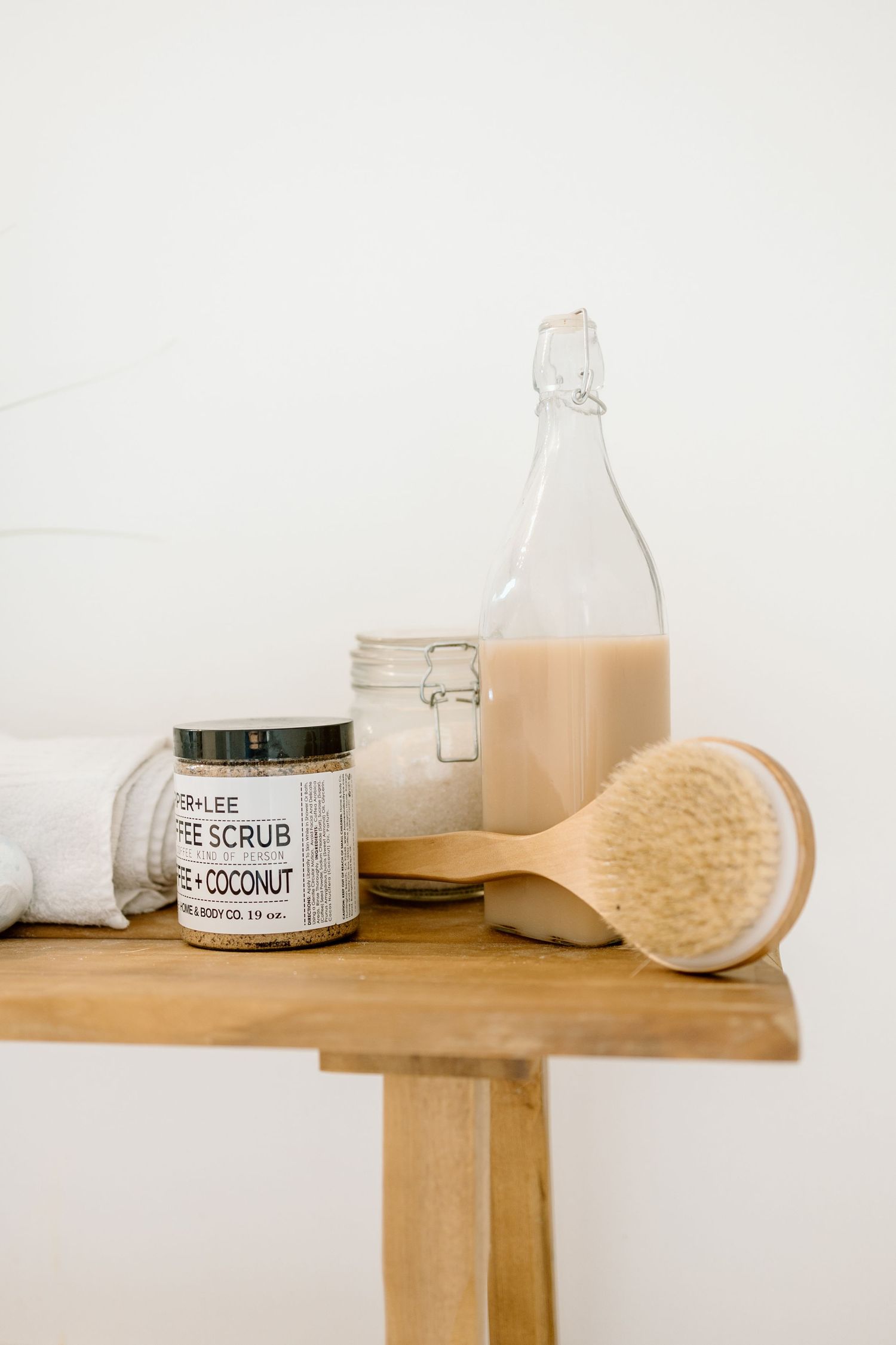 Wooden stool with coffee scrub jar, glass bottle, white towel, and body brush.