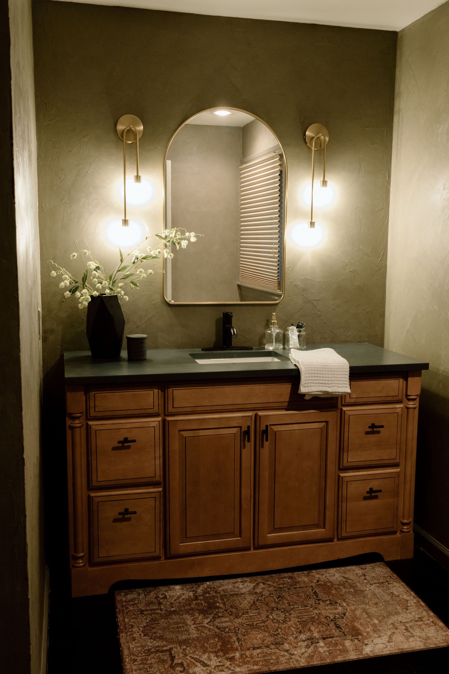 Warm bathroom vanity with wood cabinets, dark countertop, arched mirror, and brass wall sconces.