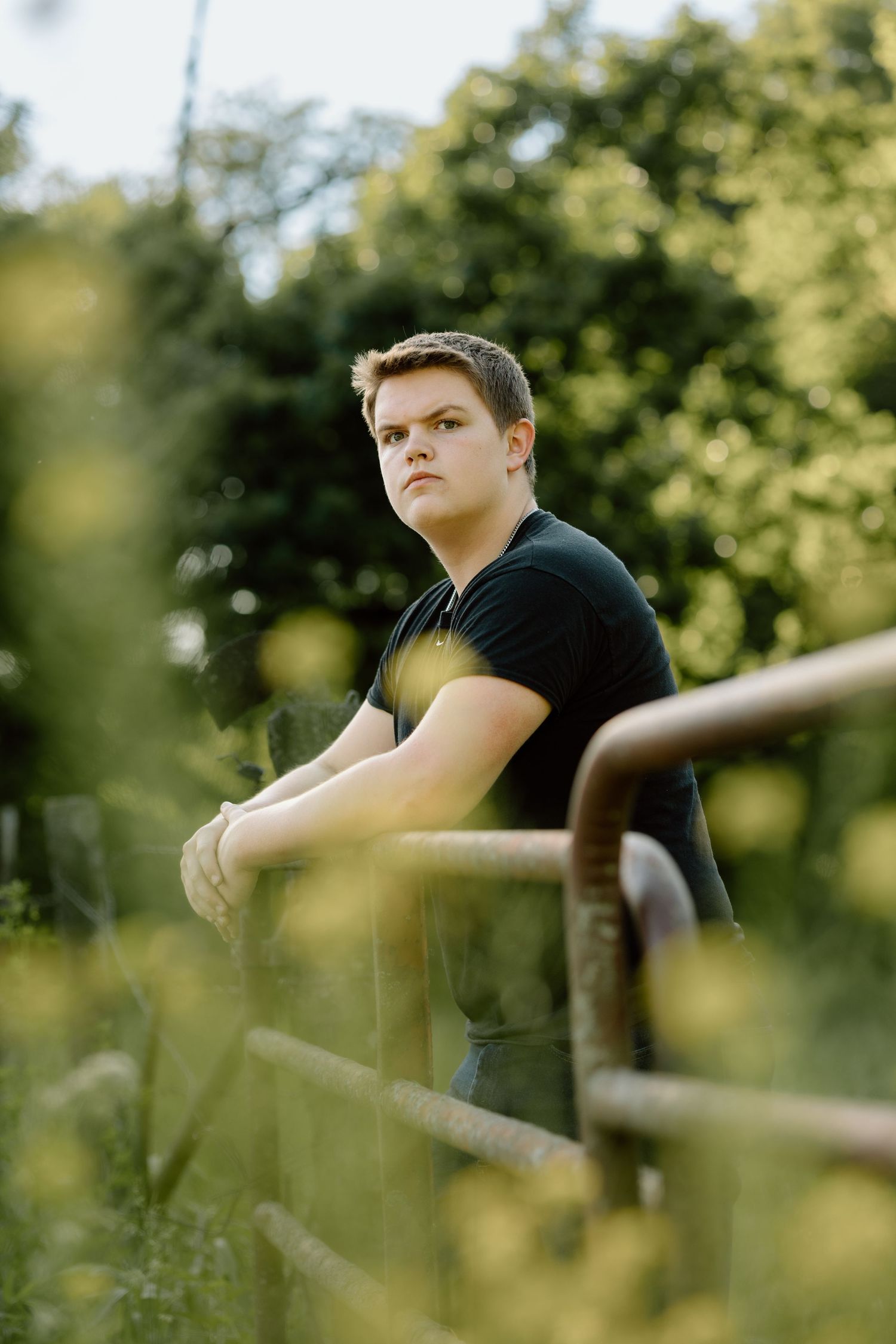 Young man in black shirt leaning on rustic fence surrounded by wildflowers in golden sunlight.