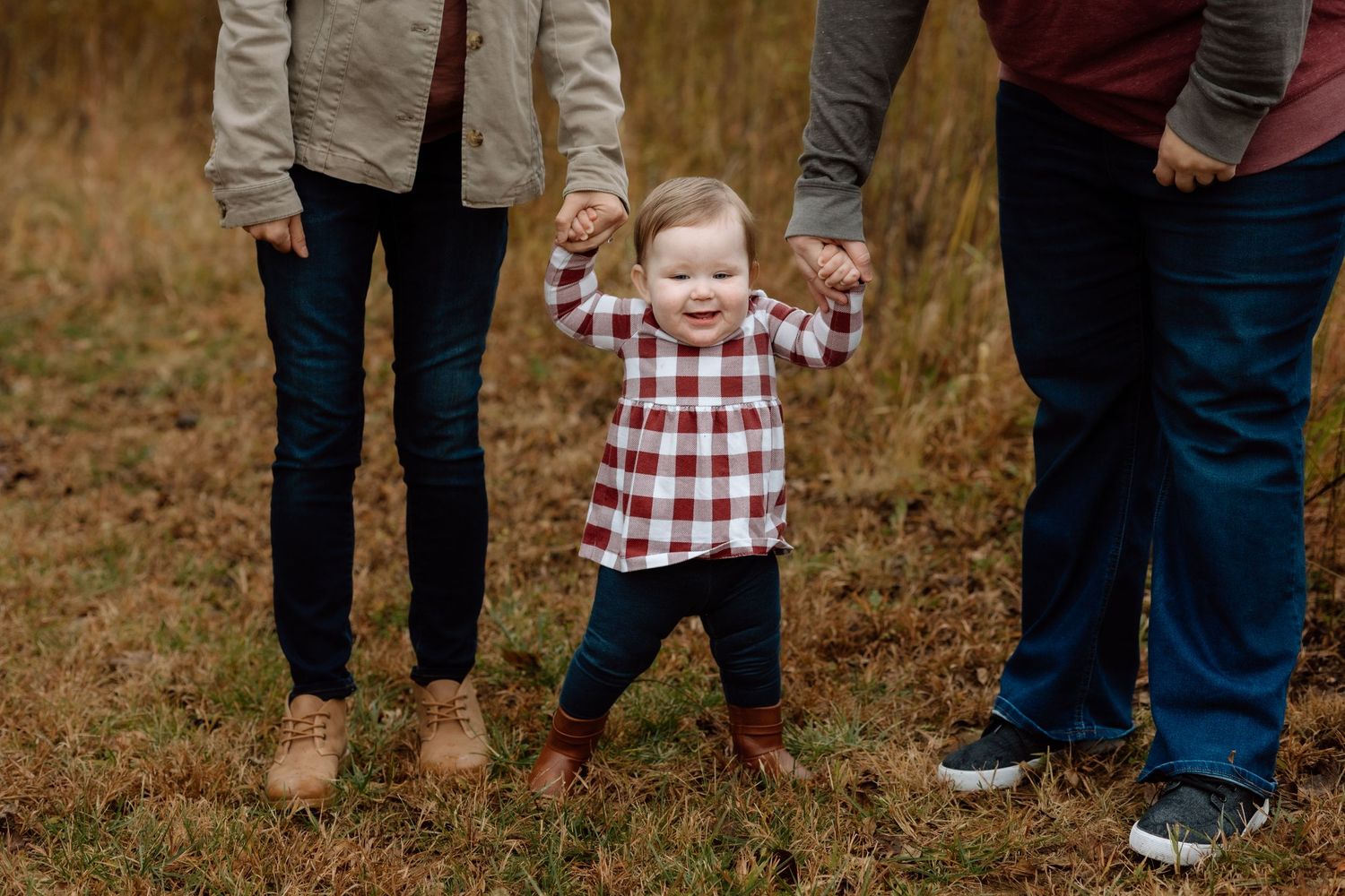 Adorable toddler in plaid dress stands holding parents hands in an autumn field during family photos.