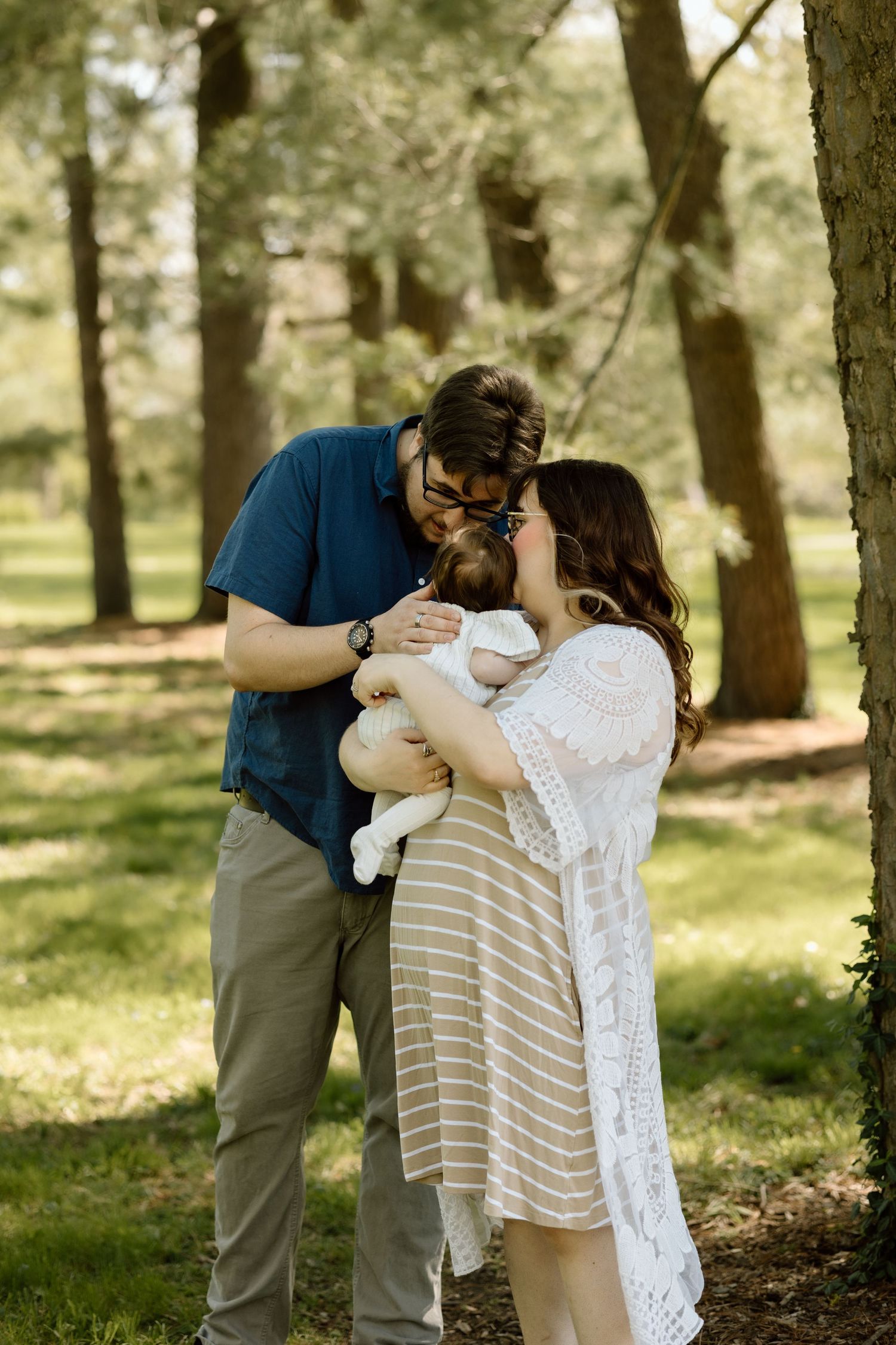 New parents lean in together to kiss their newborn baby while standing among trees in a sunlit park.