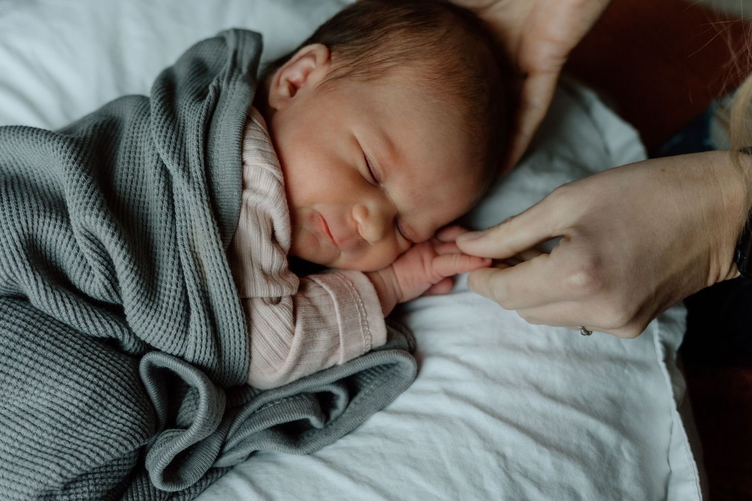 Sleeping newborn baby in gray swaddle holding adult finger gently on white blanket.