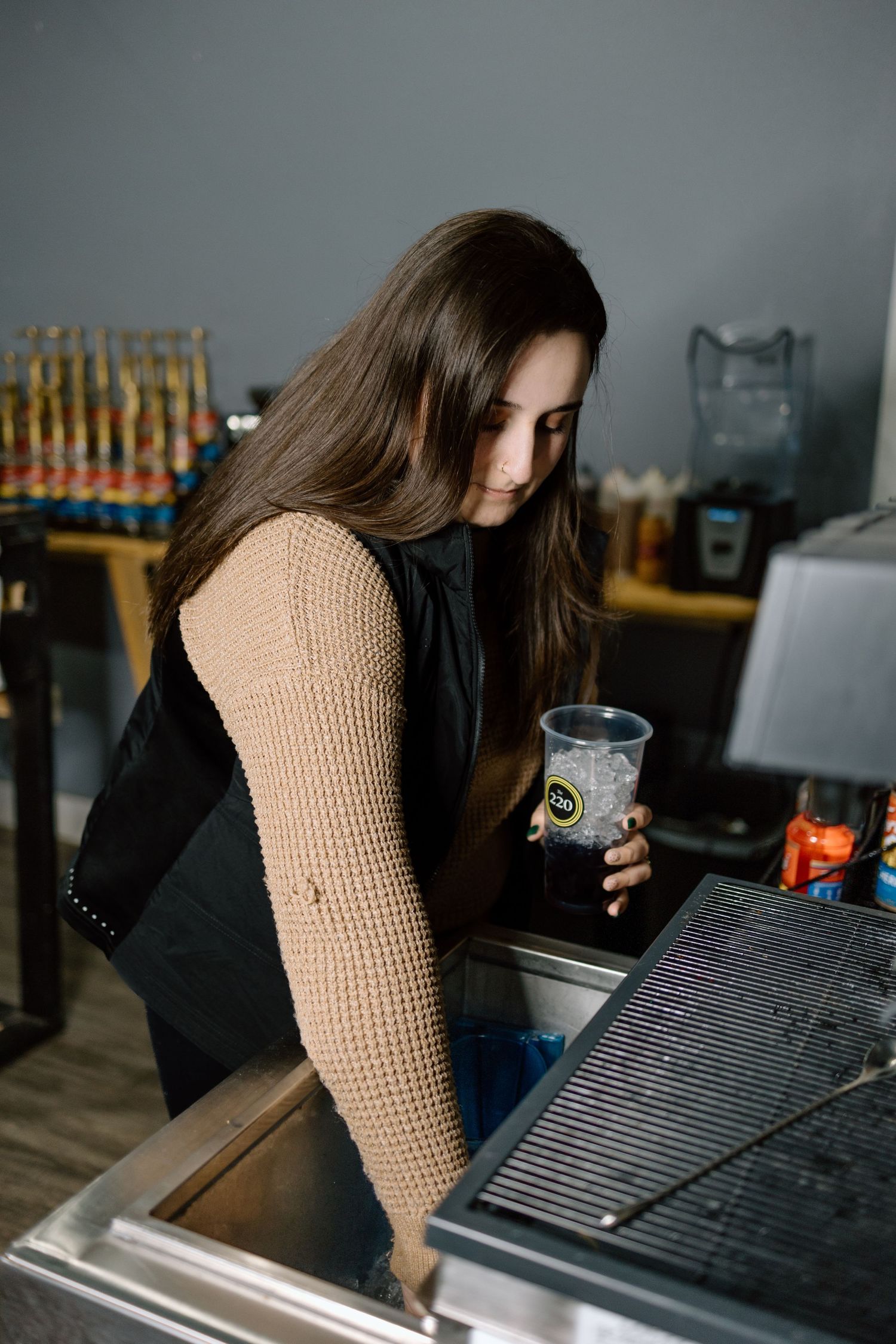 Young brunette woman in beige waffle knit jacket holding clear cup at a retail counter.