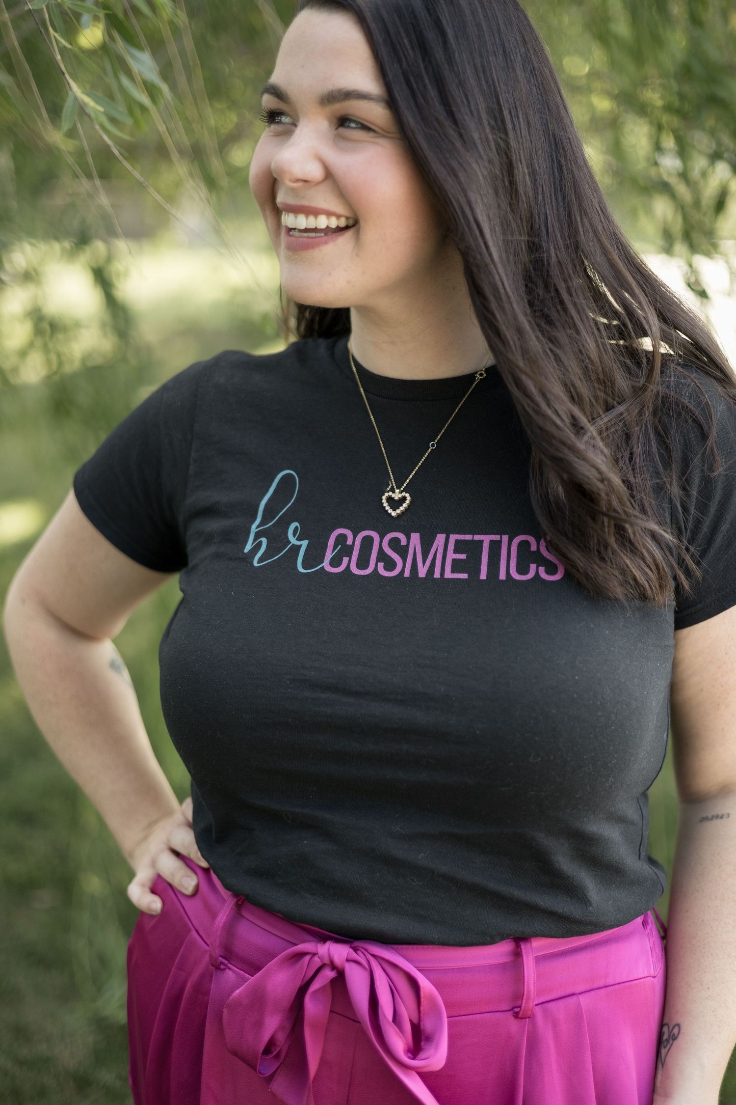 Close-up of smiling woman in HR Cosmetics black t-shirt with gold heart necklace in outdoor setting.