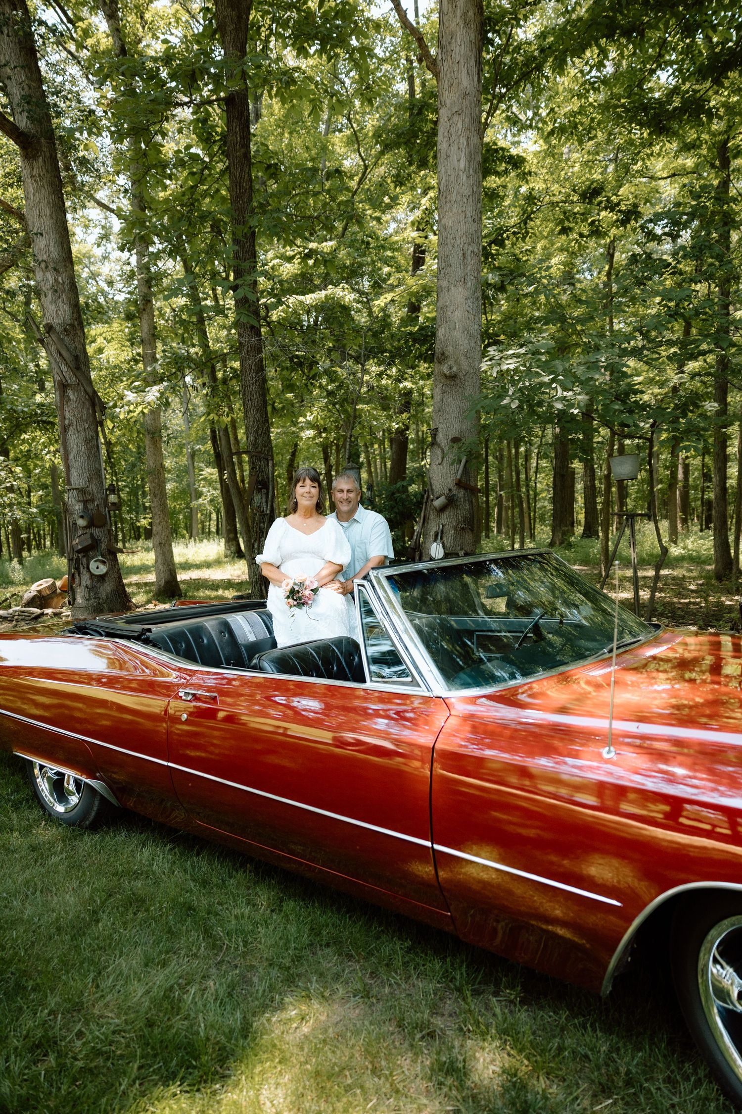Happy couple standing in red vintage Cadillac convertible convertible surrounded by lush forest trees.
