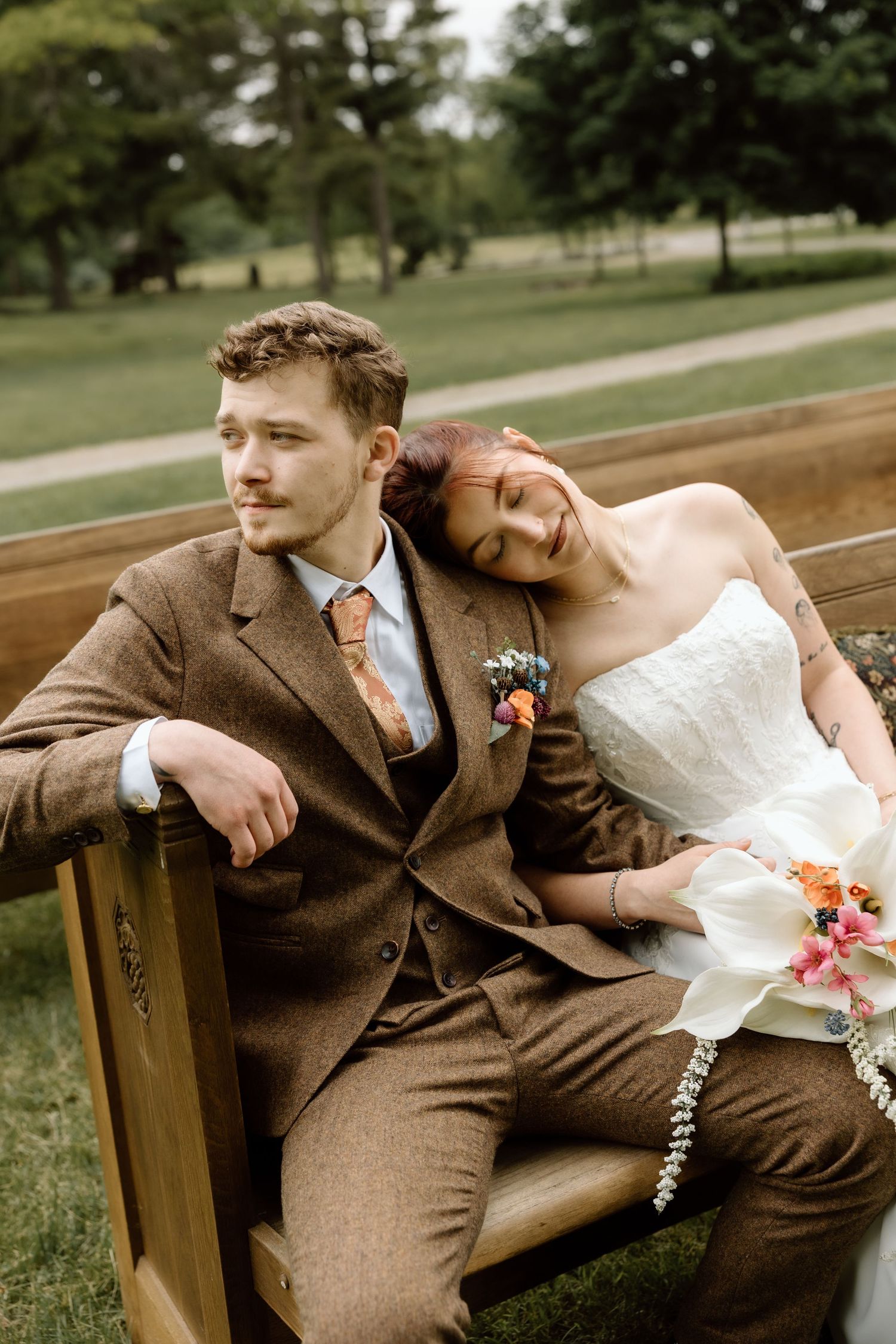 Bride in white strapless gown leaning on groom in brown tweed suit seated on a wooden bench outdoors.