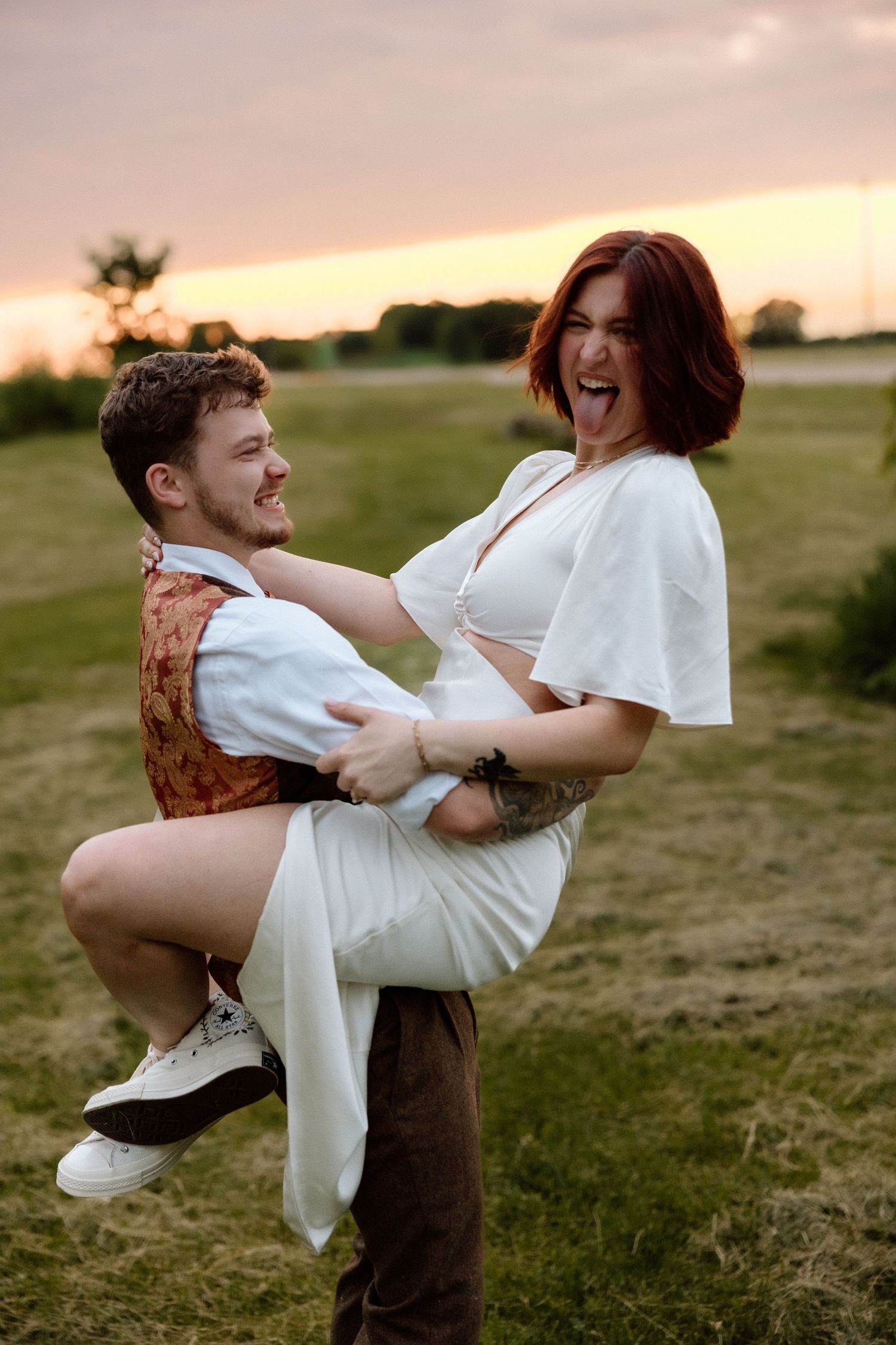 Joyful couple at sunset, man lifting laughing woman in white dress in a green field.