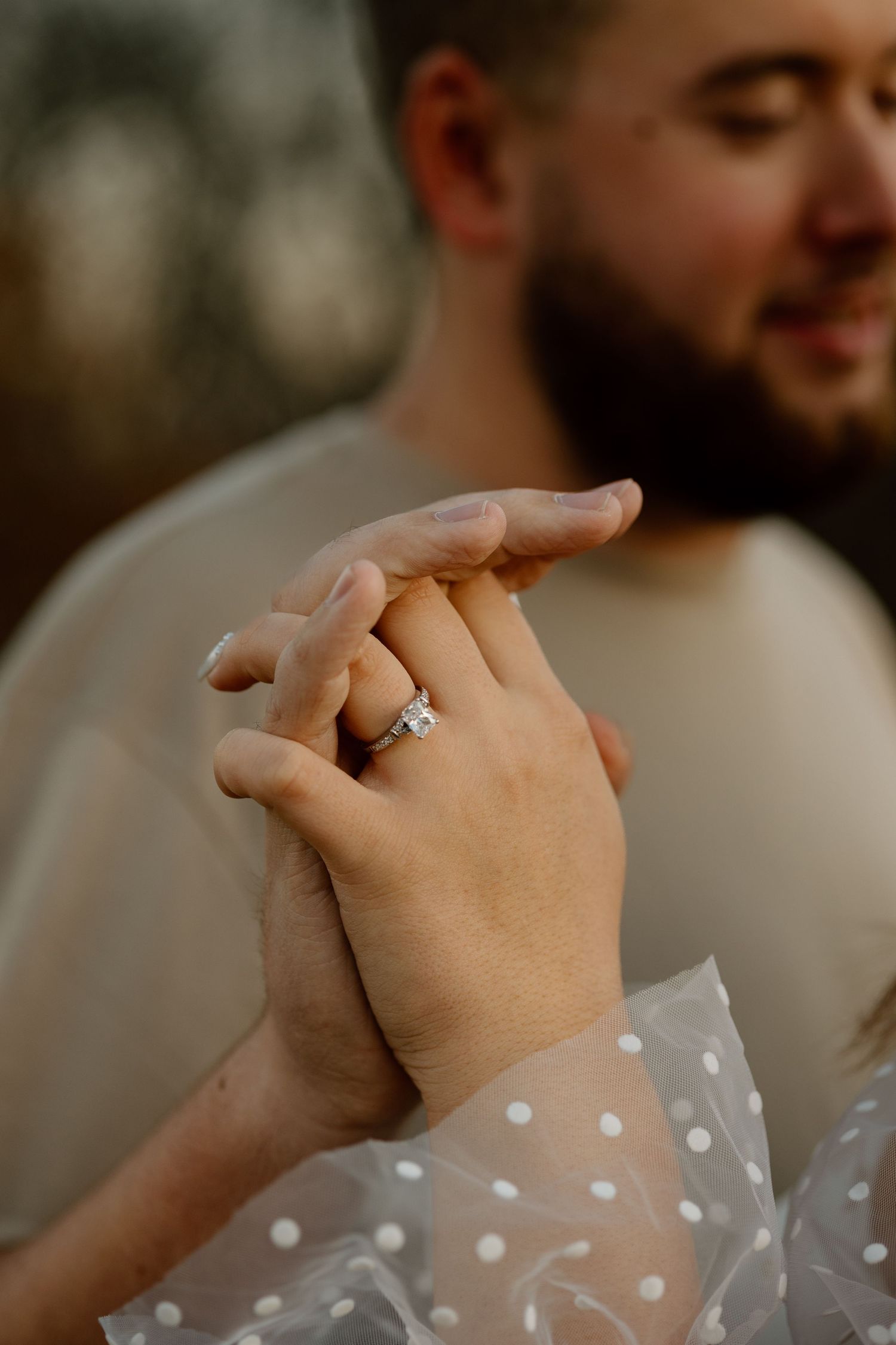 Close-up of couple holding hands showing engagement ring with sheer polka dot sleeve detail in soft focus.