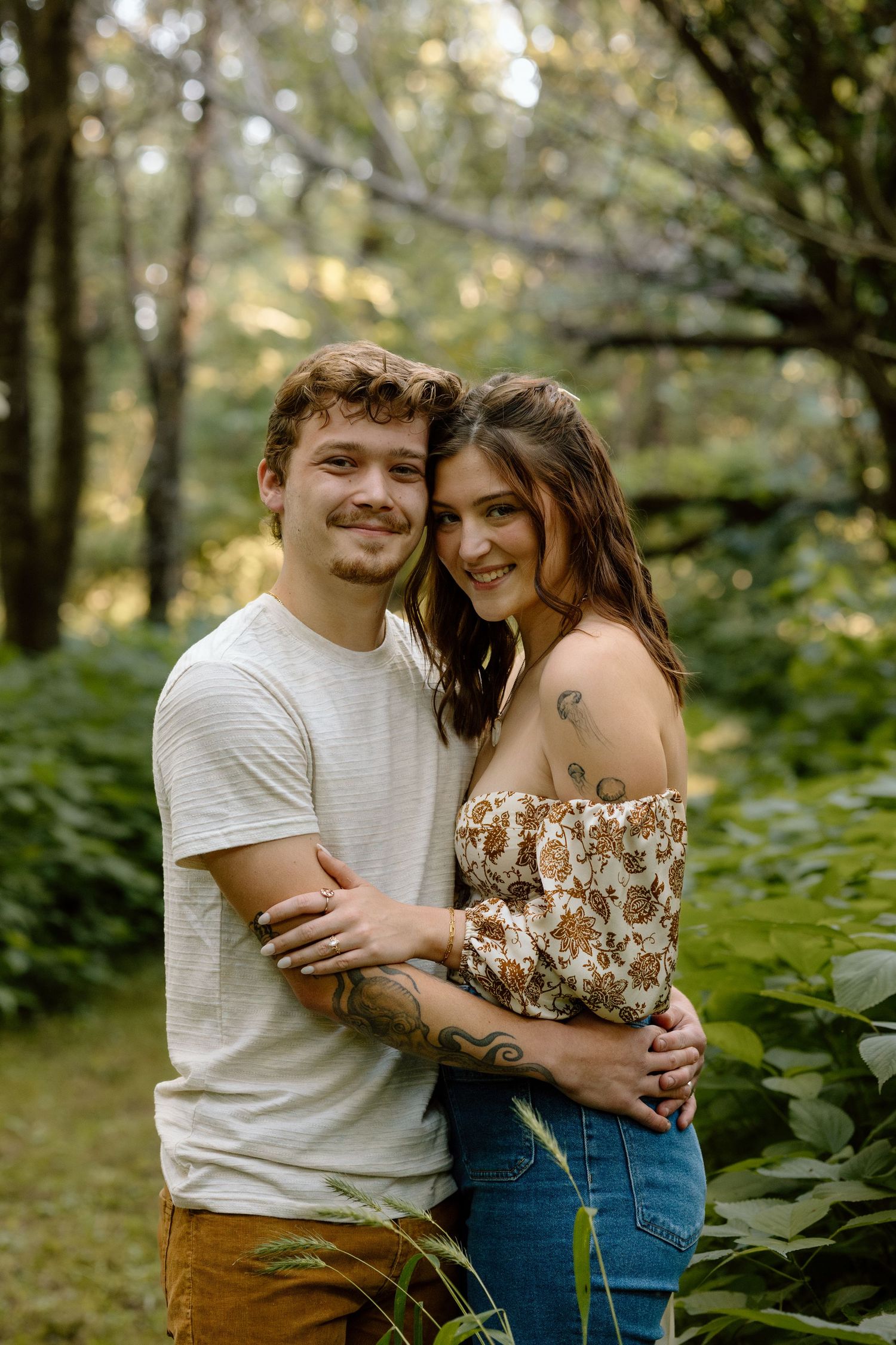 Smiling couple embracing in a sunlit forest, woman wearing floral off-shoulder top with tattoos.